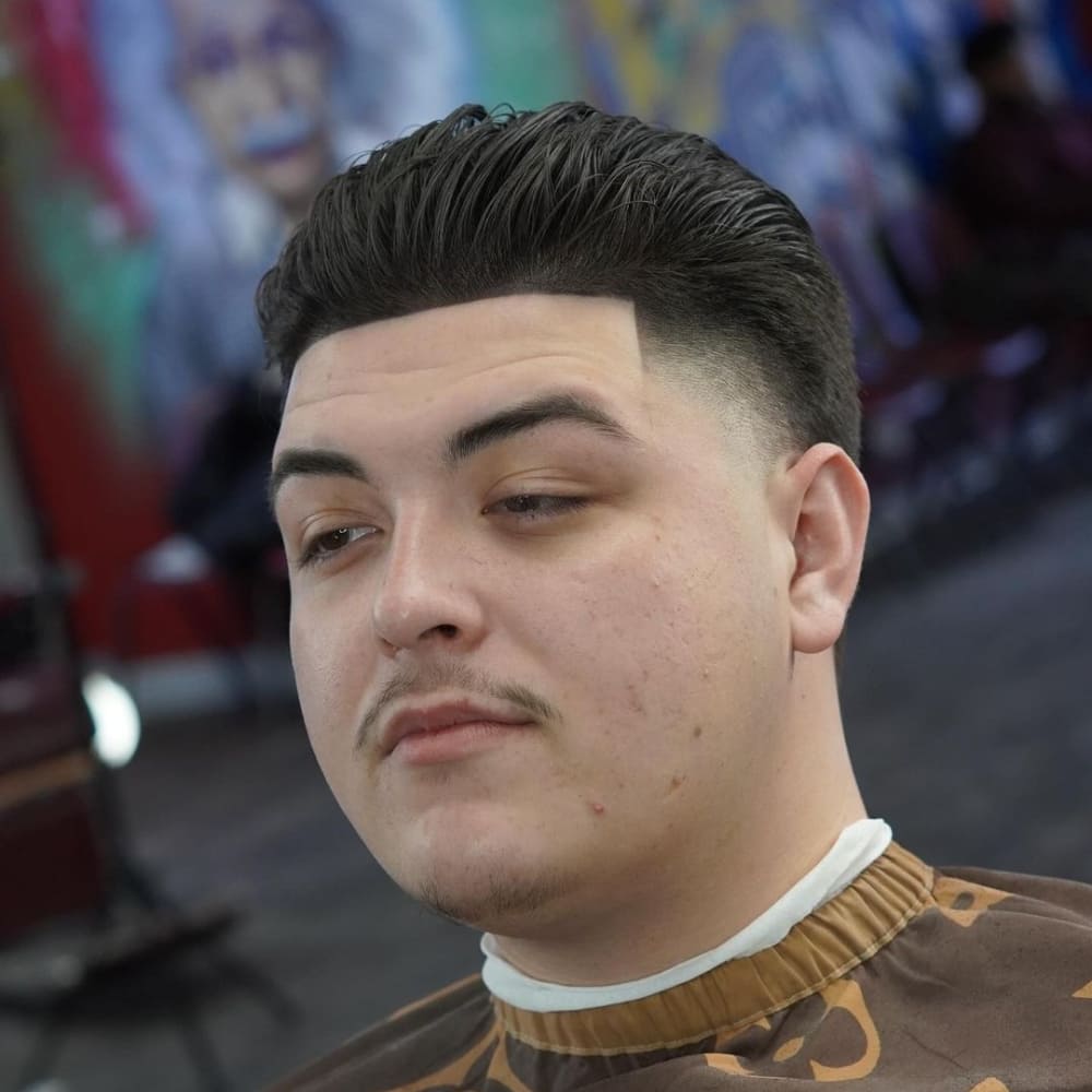 Man with a high taper haircut featuring a slicked-back top. The style showcases neatly tapered sides and a clean, defined hairline, complemented by a trimmed mustache and goatee.