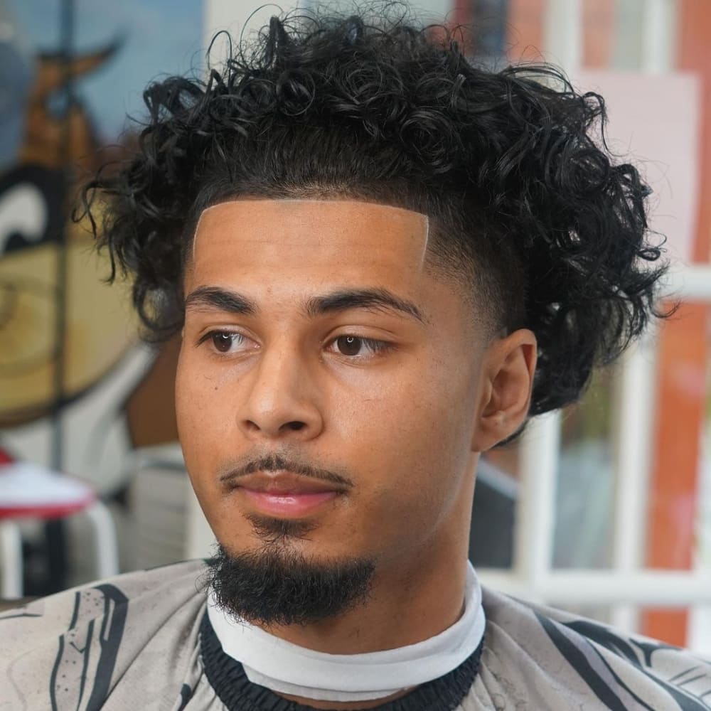 Man with a high taper undercut haircut, featuring voluminous curly hair on top and sharply tapered sides. The style showcases a clean, defined hairline and a neatly trimmed goatee.