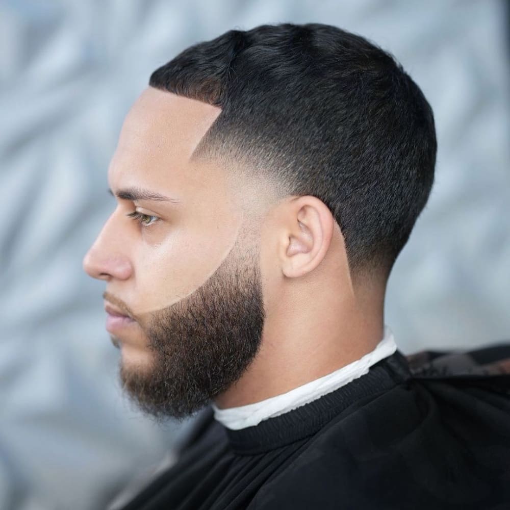 Man with a high taper haircut featuring a well-groomed full beard. The style showcases sharply tapered sides and a clean, defined hairline, creating a cohesive and polished look.