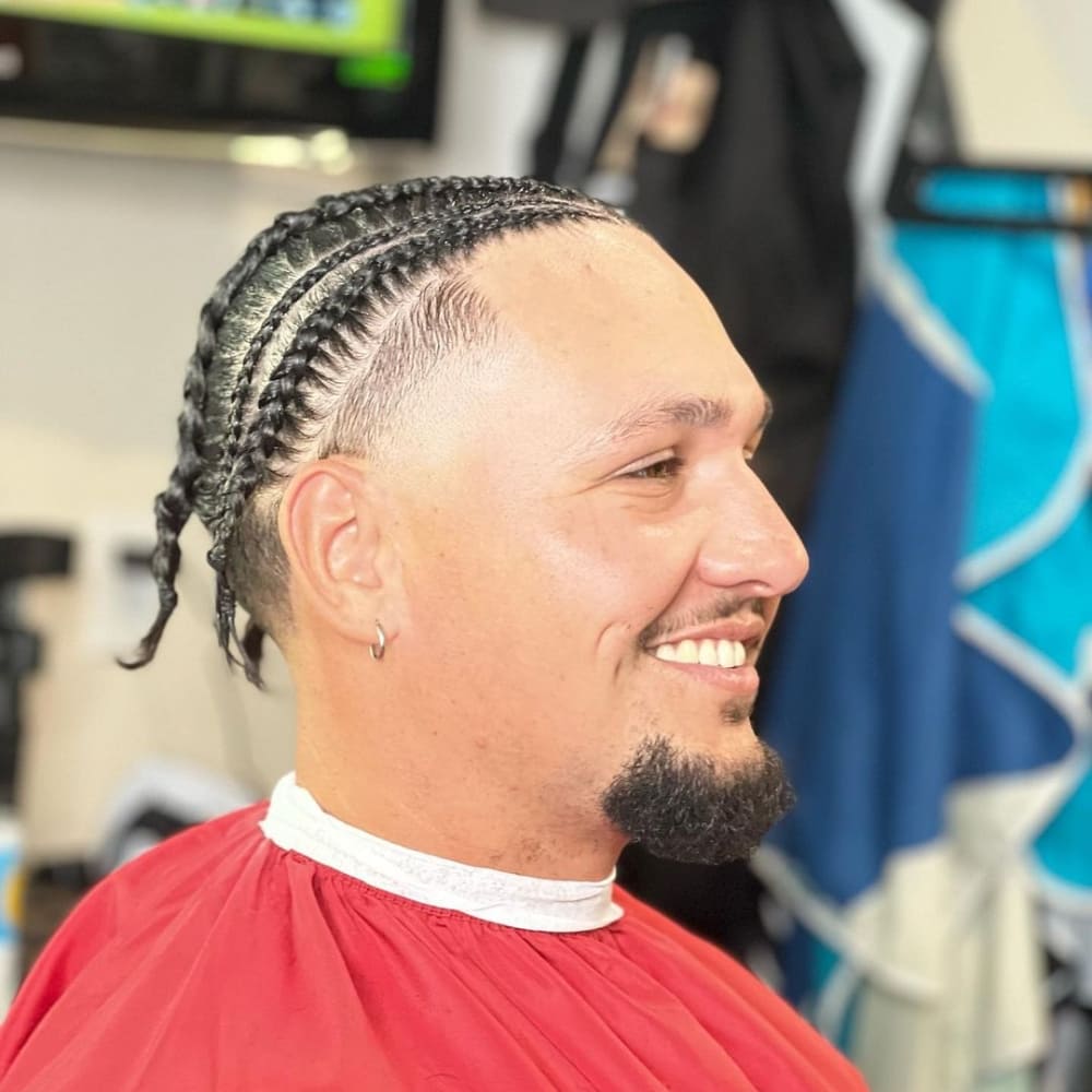 Man with a high taper haircut featuring neatly braided hair on top, showcasing cornrows that extend towards the back. The style highlights the sharp taper on the sides and a well-groomed goatee, complemented by a cheerful smile.