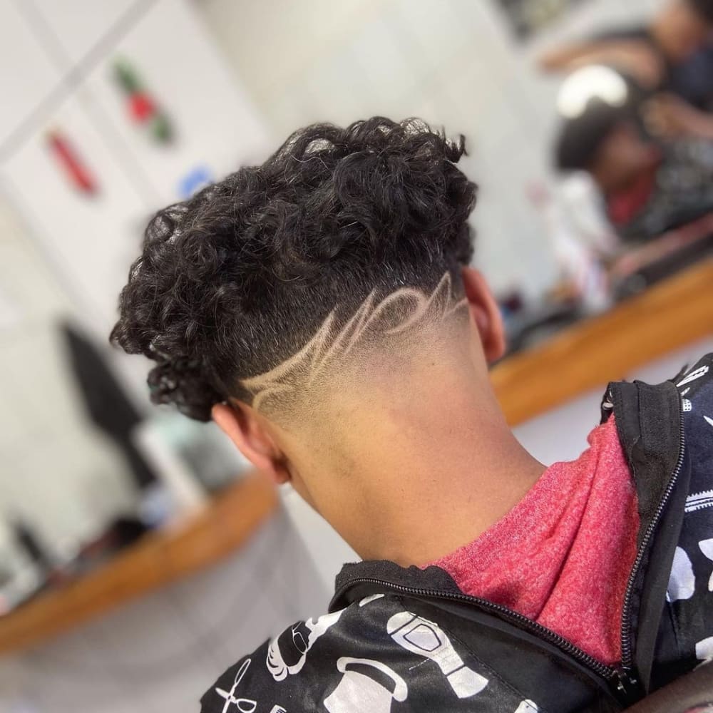 Man with a high taper haircut featuring intricate design shaved into the tapered sides and back. The style showcases voluminous curls on top, creating a striking contrast with the artistic pattern below.