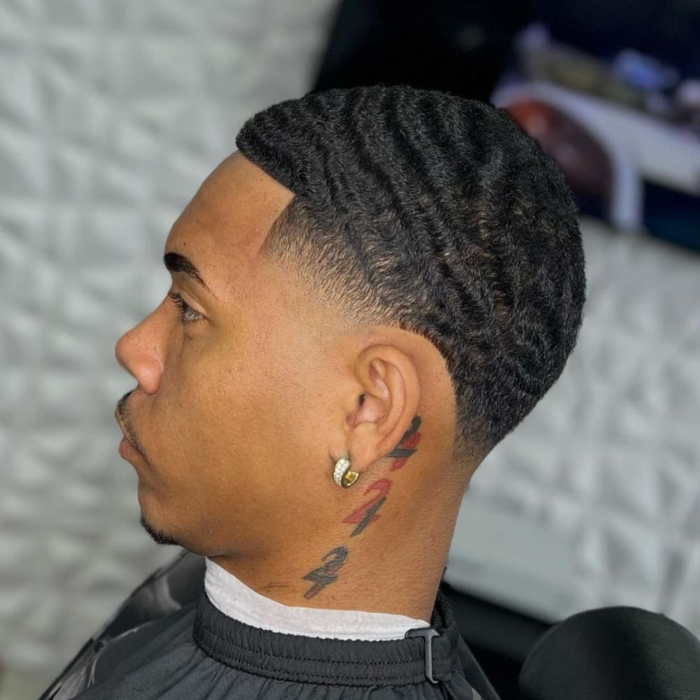 Man with a high taper haircut featuring natural waves, neatly tapered sides, and a clean neckline. The style highlights the wave pattern on top, complemented by an ear piercing and a colorful neck tattoo.