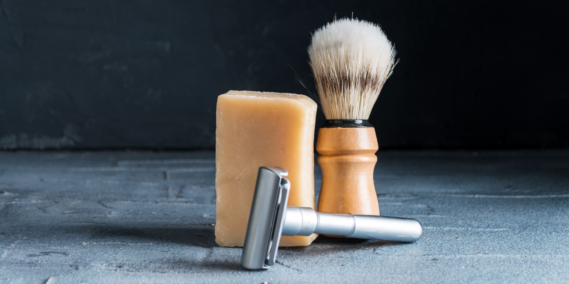 Shaving brush with wooden handle, bar of soap, and safety razor on a gray surface. Learn how to clean a shaving brush, including badger hair brushes, for a hygienic and effective grooming routine.