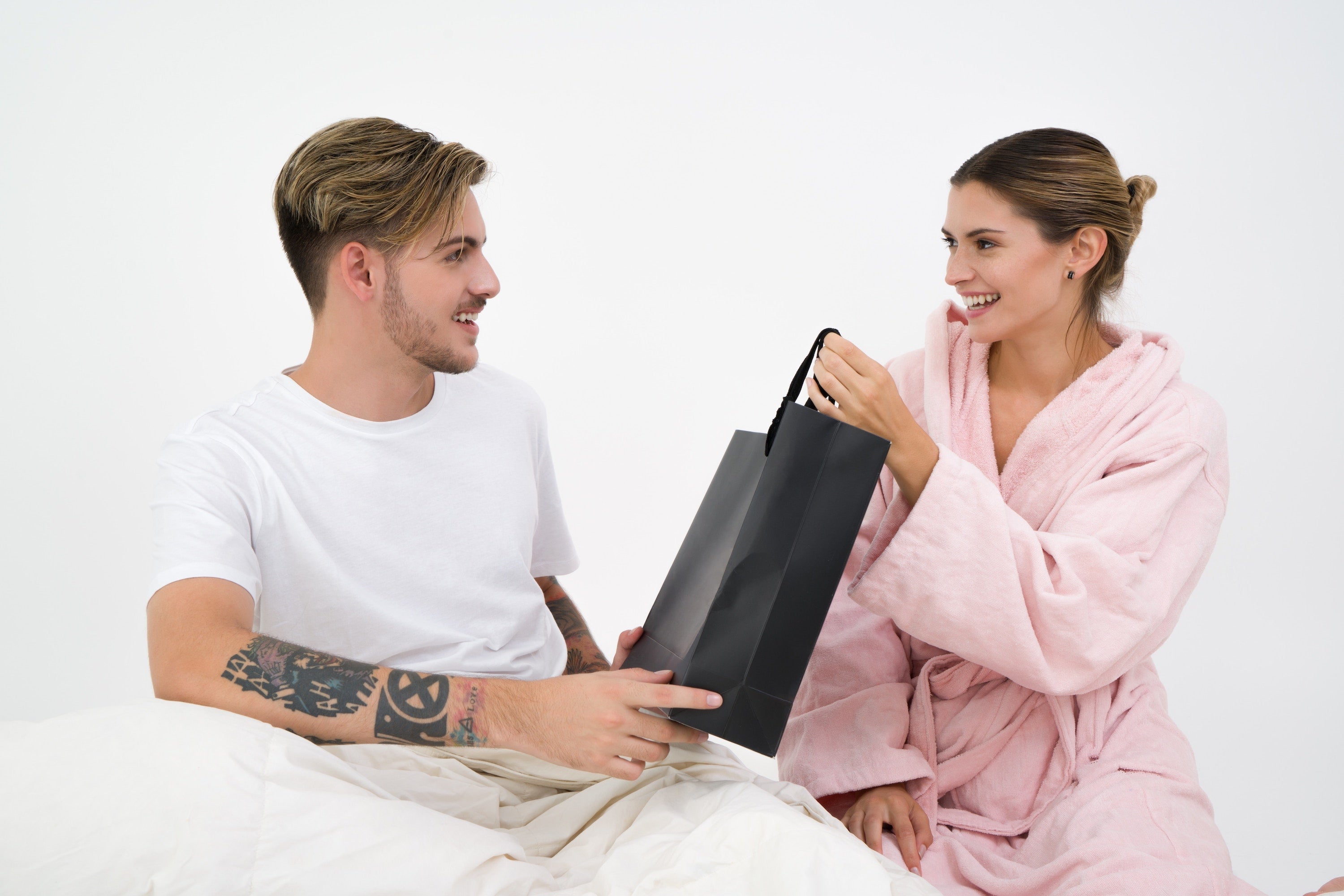 A woman in a pink robe is smiling and handing over a black shopping bag to a man in a white t-shirt who is sitting on a bed. Both are looking at each other with joyful expressions, suggesting the exchange of a gift. The man has visible tattoos on his arms