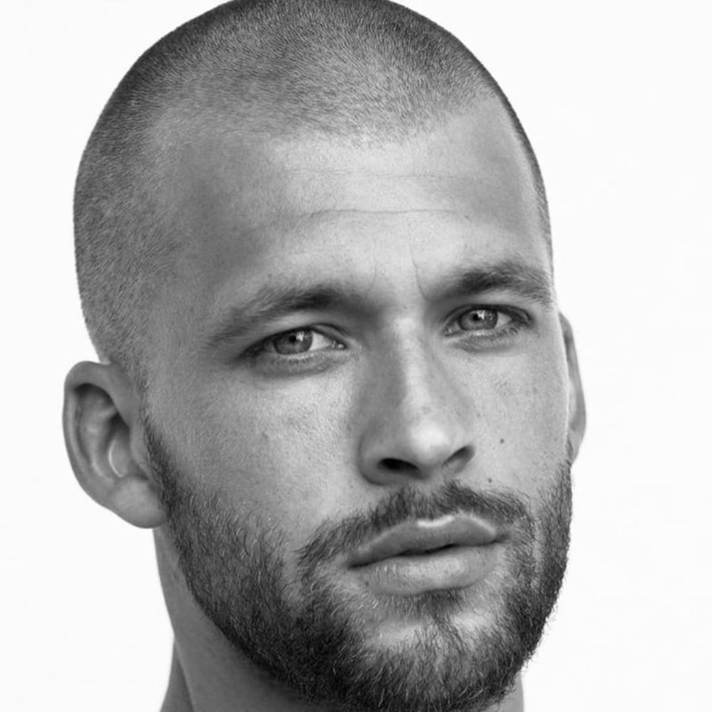 A close-up, black-and-white portrait of a man with an induction buzz cut. The haircut is extremely short, with the hair clipped very close to the scalp, giving a nearly bald appearance. The man’s facial features are emphasized by the simplicity of the haircut, showcasing a strong jawline and neatly groomed beard that adds to the rugged, masculine look. His expression is serious, highlighting the bold and confident nature of the induction buzz cut. The overall style is minimalistic, clean, and traditionally associated with military grooming.