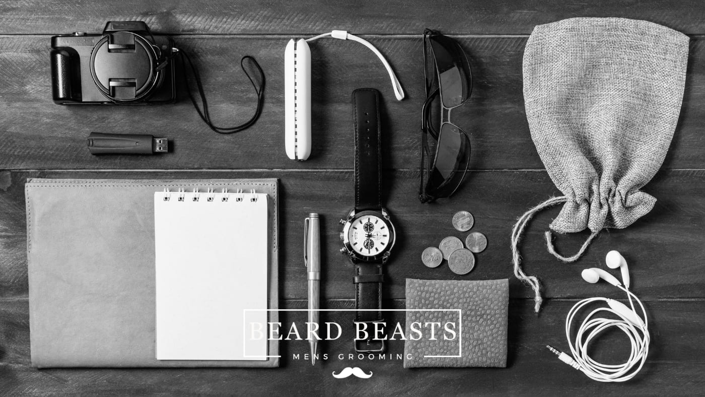 Assorted collection of essential items for men, featuring a camera, USB drive, toothbrush, notebook, pen, watch, sunglasses, loose change, drawstring bag, and earphones, all arranged on a wooden background, labeled with Beard Beasts Men's Grooming.