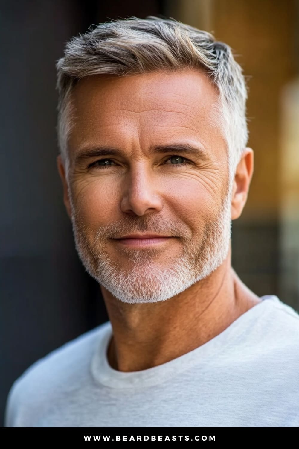 A confident older man with a well-trimmed beard and a stylish haircut that exudes timeless charm. His hair is neatly cut with slightly longer strands on top, combed over to create a polished and refined look. The sides are trimmed shorter, blending seamlessly into the top, giving his overall appearance a balanced and sophisticated edge. The soft lighting and neutral background enhance his natural features, capturing a look that is both modern and effortlessly elegant.