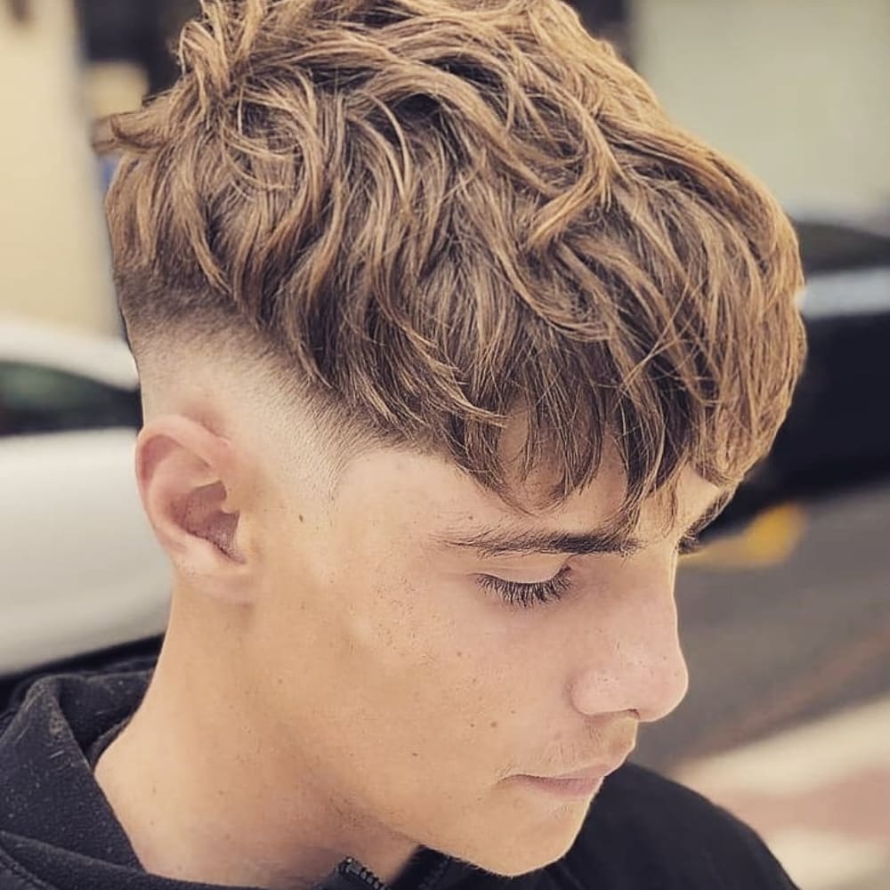 Young man with a long Caesar haircut featuring textured, wavy hair on top that extends down to a straight fringe. The sides and back are faded, creating a sharp contrast with the longer top. He is wearing a black hoodie and looking down, showcasing the stylish and voluminous top.