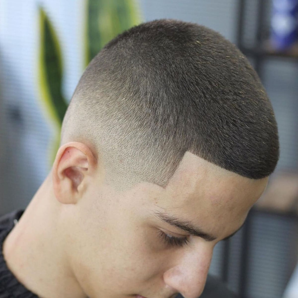 A close-up profile of a young man with a neatly trimmed, short haircut featuring precise, clean lines and a smooth gradient on the sides. The hairstyle is expertly detailed, emphasizing sharp edges and a polished look. The soft lighting and subtle background elements, such as a blurred plant, highlight the sleek simplicity of the cut while maintaining a fresh and modern aesthetic.