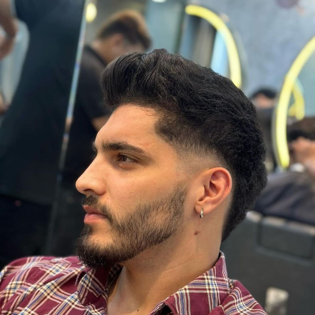 Side profile of a low burst fade quiff hairstyle featuring a rounded fade around the ears and a high-volume, textured top. This low burst fade quiff is ideal for adding a modern, edgy look with natural waves and a well-groomed beard for added style."