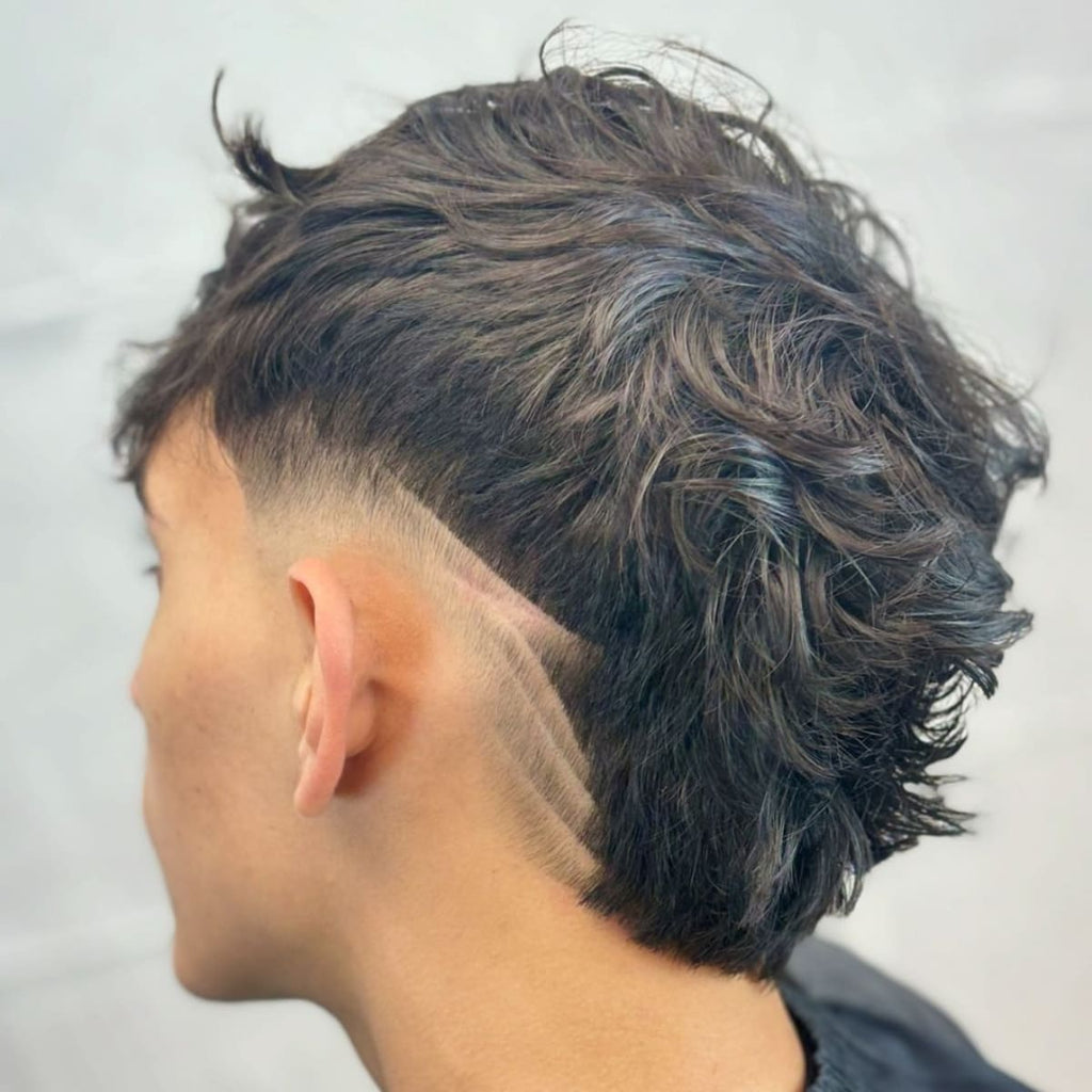 Side view of a stylish low burst fade mohawk haircut featuring wavy textured hair on top and sharp, geometric line designs shaved into the fade. The clean, edgy look highlights the contrast between the longer hair and the detailed fade design.