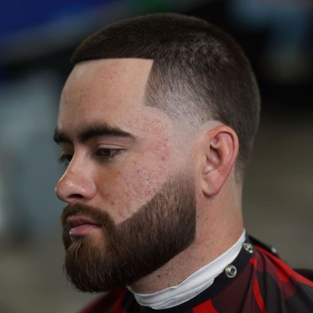 A close-up side profile of a man with a low fade buzz cut combined with a line-up and a full beard. The haircut features short, even hair on top with a gradual fade down the sides, blending smoothly into the skin. The hairline is sharply defined with a precise, straight line-up at the forehead and temples, creating a crisp and clean look. The man’s beard is thick and meticulously groomed, with sharp edges that align perfectly with the haircut, enhancing the overall structured and polished appearance. The style is modern and stylish, ideal for those who appreciate sharp lines and a well-defined look.