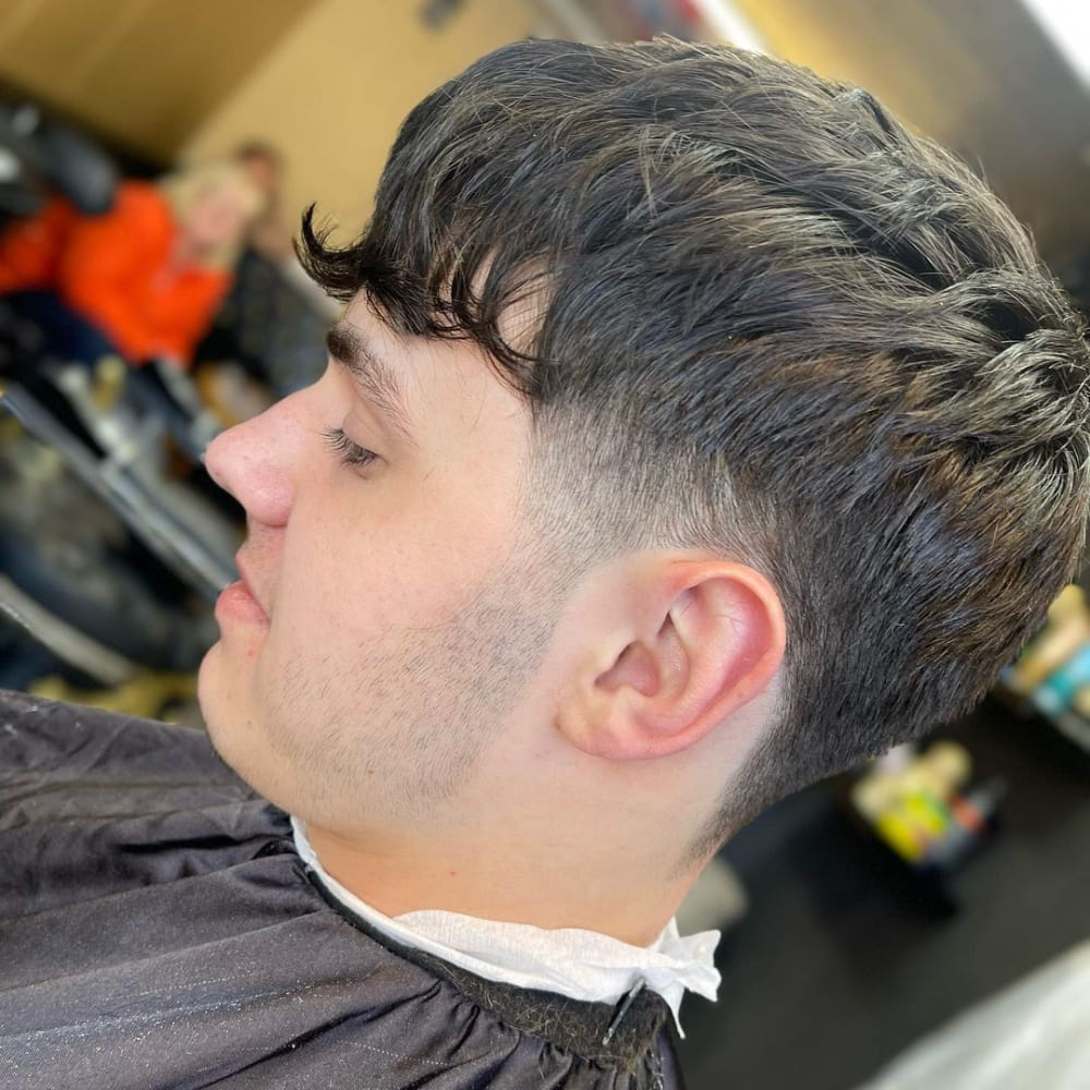 Young man with a Caesar haircut featuring a wavy, slightly tousled fringe and a low fade on the sides and back. The textured top adds volume and movement, while the low fade provides a subtle transition. He is seated in a barber chair, wearing a black cape, with a blurred background of a busy barbershop.