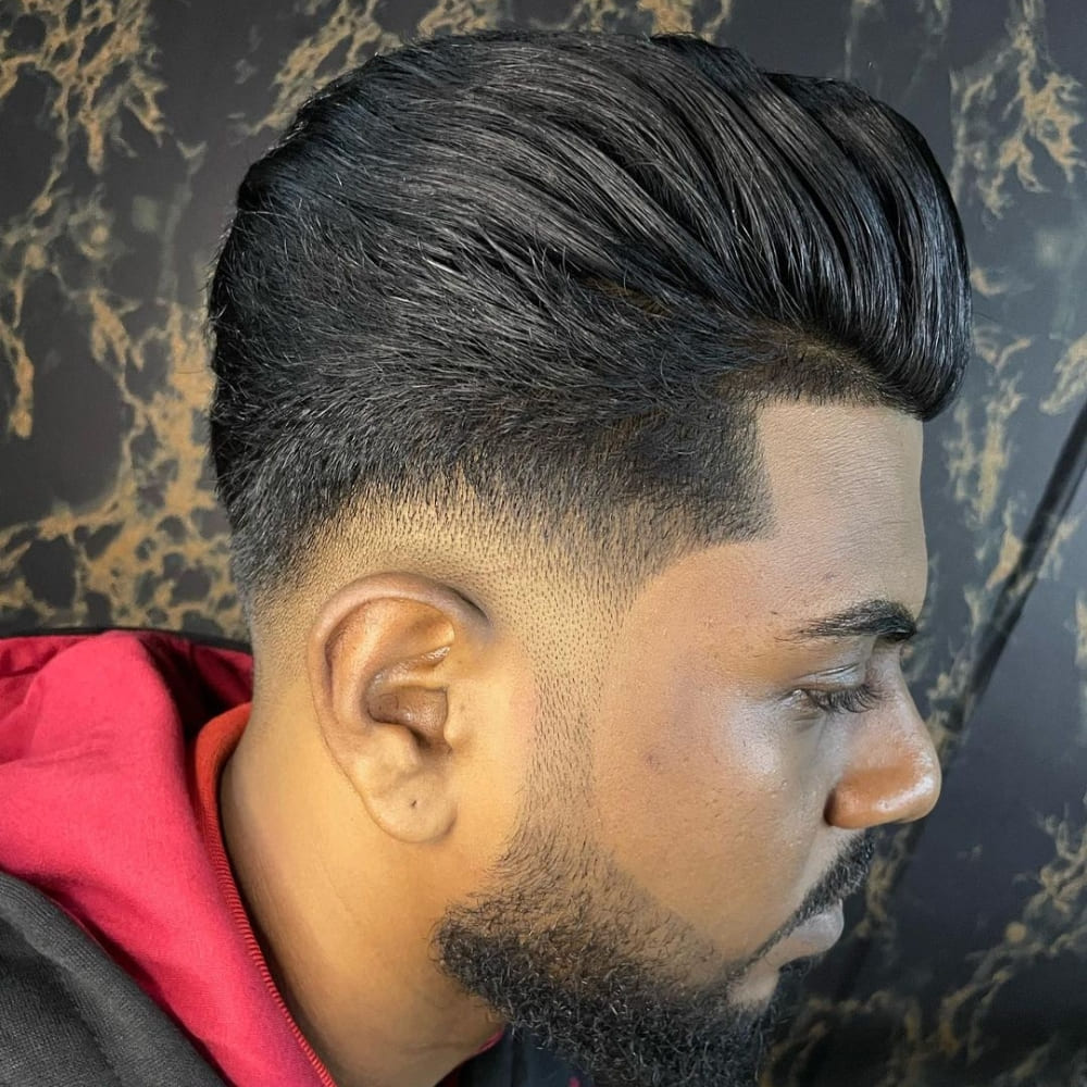 A man is showcasing a low fade pompadour haircut, which features a smooth transition from short, faded sides to a voluminous top. His thick, dark hair is styled upwards and backwards, creating a sleek and modern pompadour look. The beard is meticulously groomed, aligning perfectly with the sharp lines of the haircut. The background is a stylish marble-patterned wall, adding to the sophisticated aesthetic of the overall look. The man's side profile highlights the precision and craftsmanship of the haircut, emphasizing its clean and polished appearance.