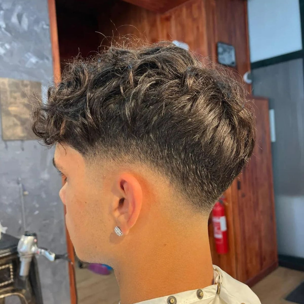 A close-up side view of a young man with a low fade haircut and a textured, curly top. His hair on top is full, with natural curls adding volume and movement, while the fade gradually tapers down along the sides and back, creating a smooth transition. He is wearing a small diamond earring, adding a subtle touch of style to his look. The background shows a barbershop interior with wooden paneling, adding a warm, relaxed atmosphere to the scene.