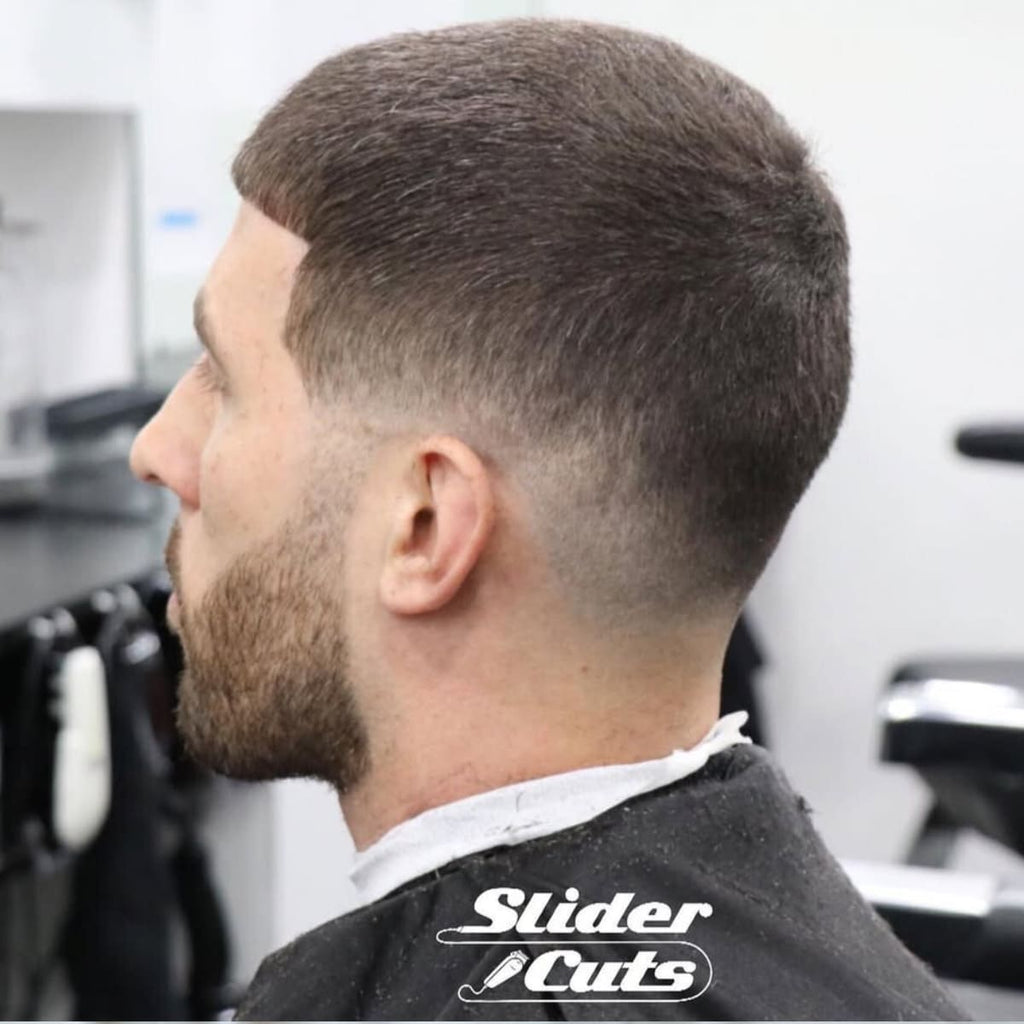 A side profile of a man with a clean haircut featuring a gradual blend that transitions smoothly from the scalp to the short, even hair on top. The style is paired with a neatly trimmed beard, creating a polished and modern appearance. The subtle blend adds a professional and understated touch to the overall look.