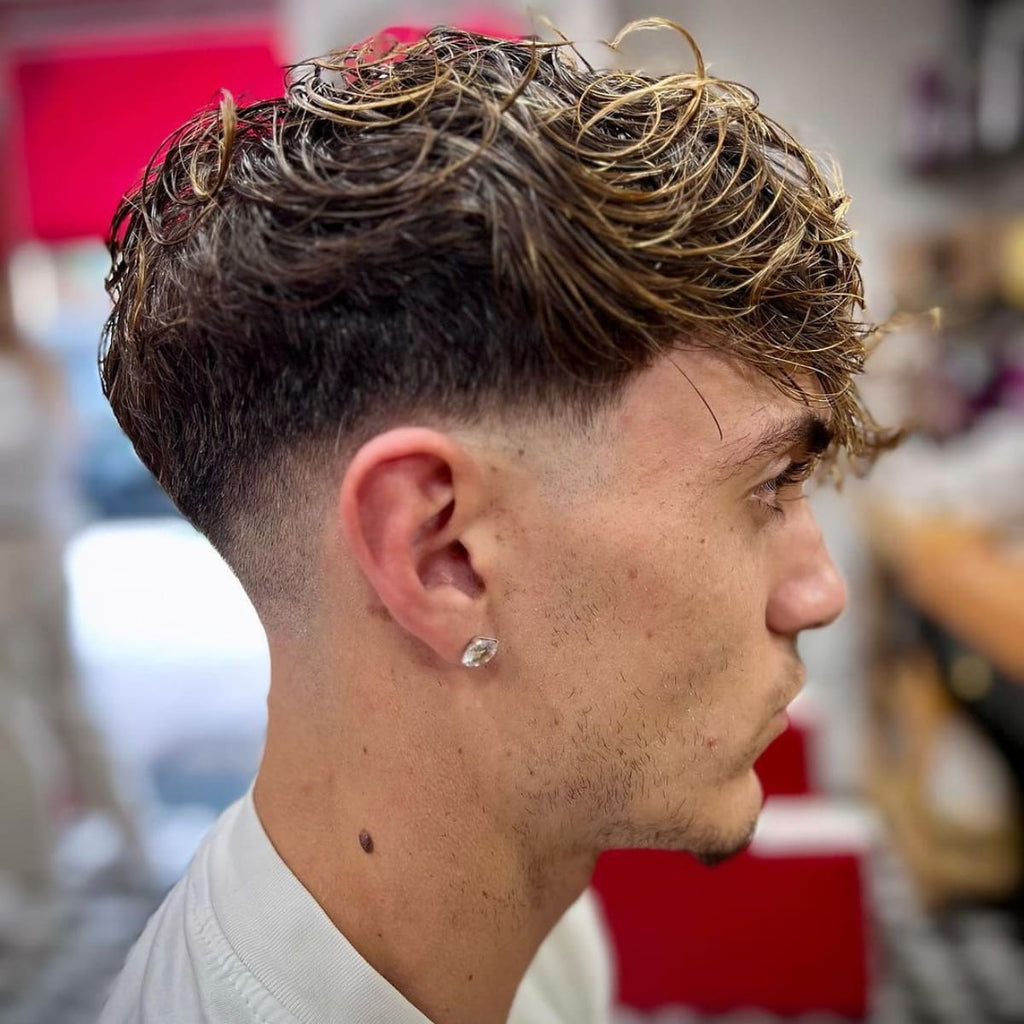 A side profile of a young man with wavy, highlighted hair styled in a loose, textured fashion on top. The hair gradually tapers down into a clean, smooth fade near the ears and back, creating a soft transition. He wears a sparkling stud earring that adds a touch of flair to his look. The background appears to be a well-lit barbershop, giving the image a casual and stylish feel.