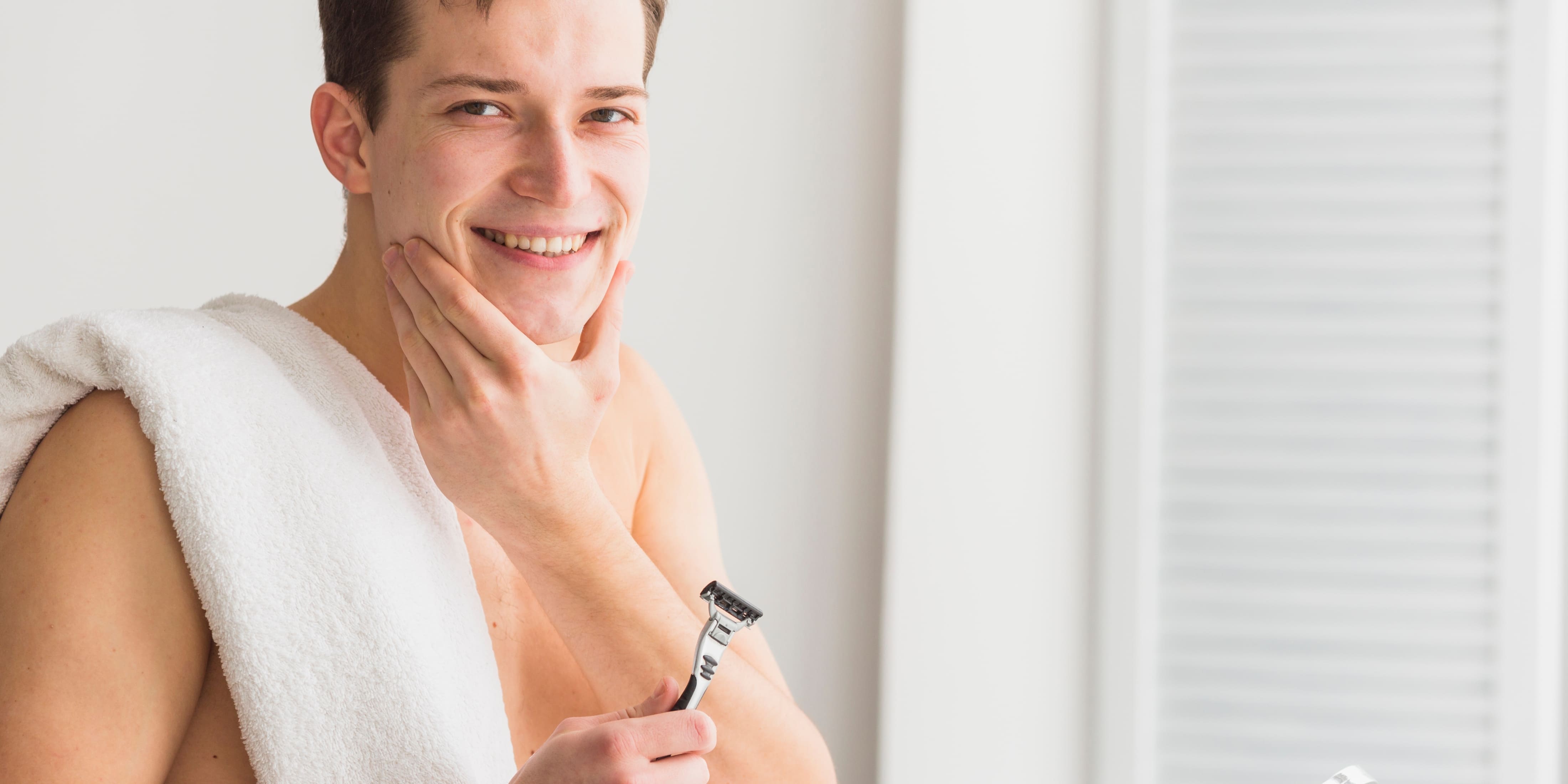 Smiling man after learning how to get a close shave, holding a razor and touching his smooth face. He has a towel draped over his shoulder and looks satisfied with his close shave. The bright, neutral background highlights his fresh, clean appearance. Keywords: how to get a close shave, close shave, smooth face, shaving.