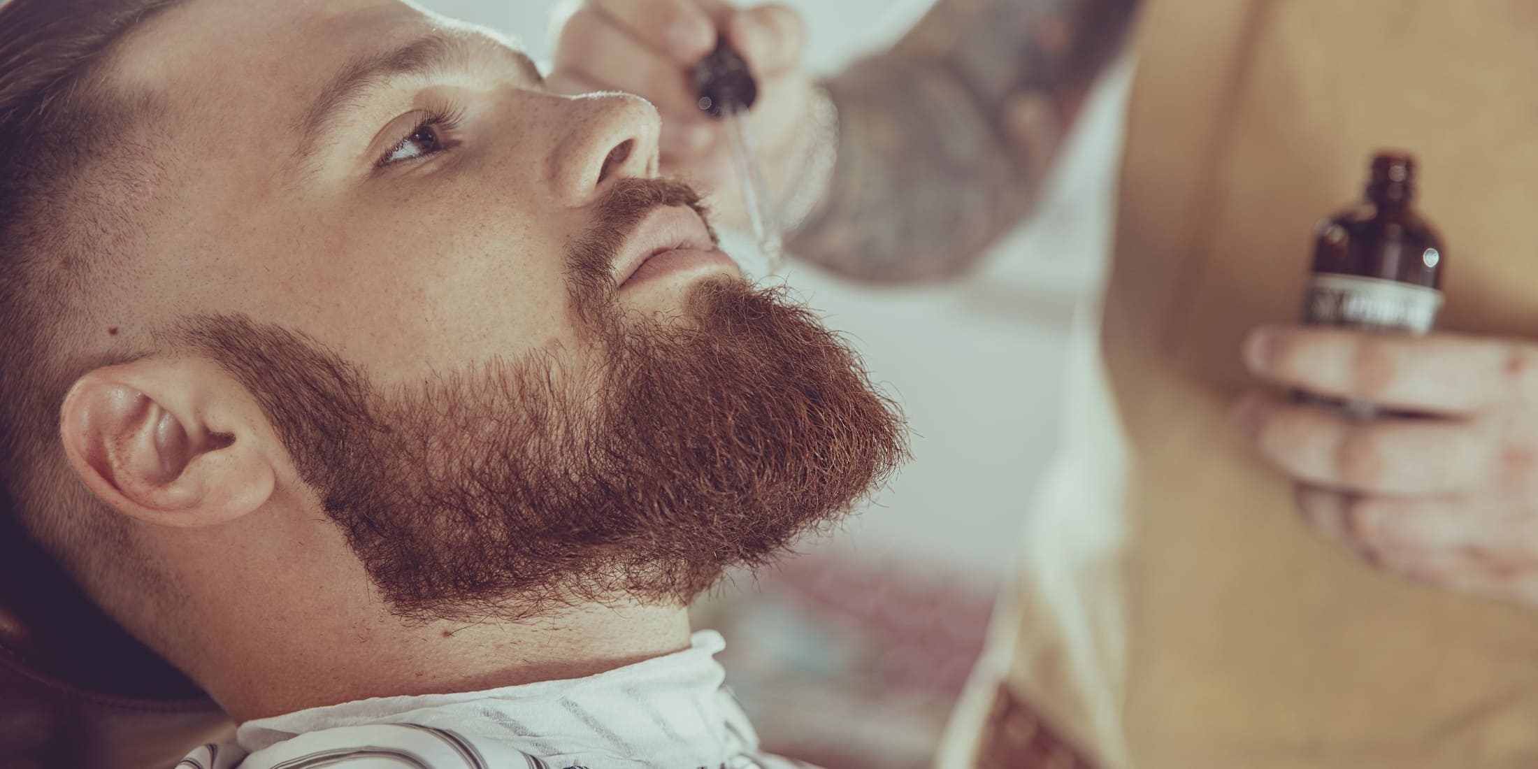 A bearded man reclining while a barber applies beard oil to his beard, a process essential for maintaining beard health and style. The barber holds a dark bottle of beard oil, ready to demonstrate what beard oil does for your beard in terms of care and grooming.