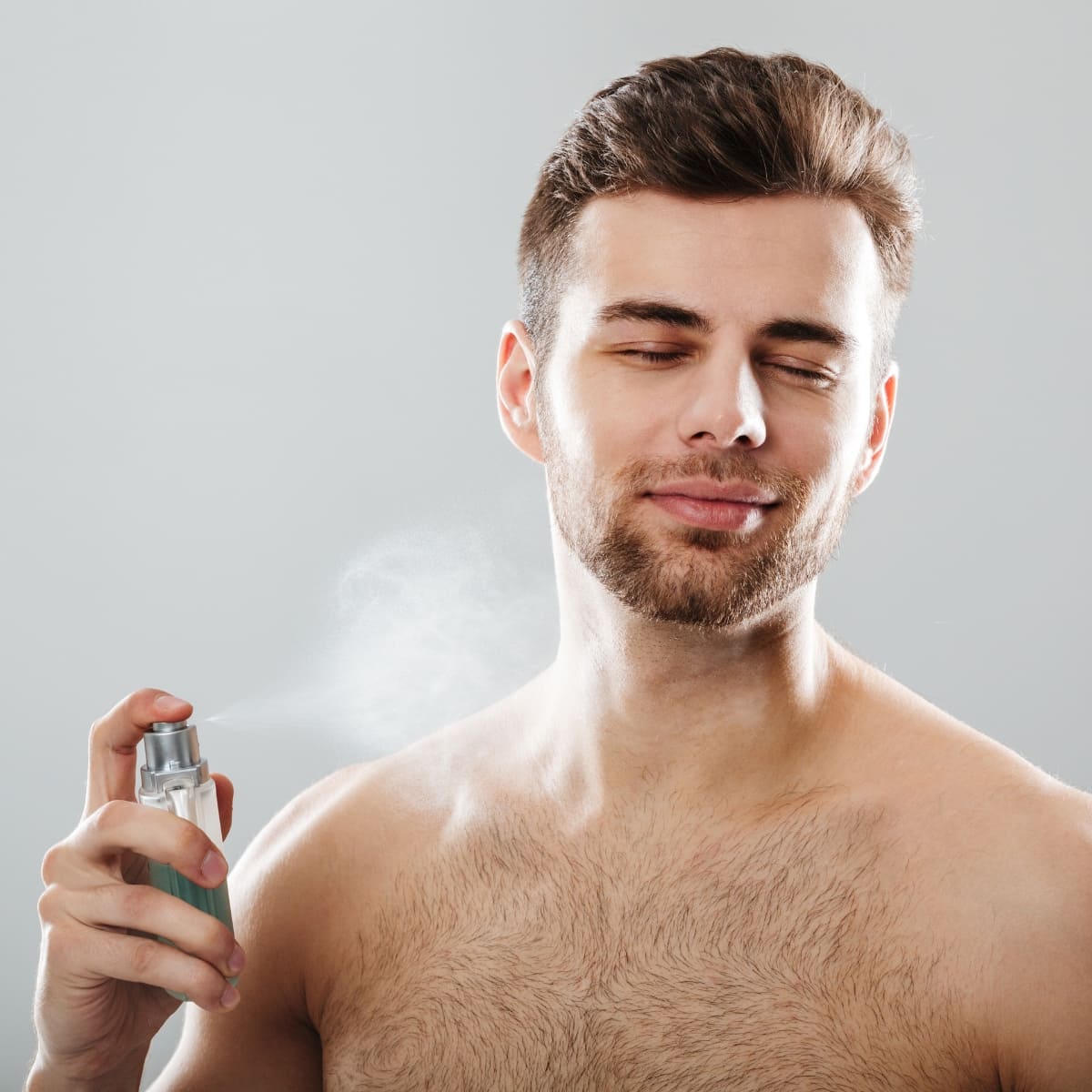 A young man with a well-groomed beard and neatly styled hair is seen applying cologne to his upper body. He is shirtless, with a slight smile on his face and his eyes closed, enjoying the fresh scent. The background is a plain, light gray color, keeping the focus on the man and his grooming routine.