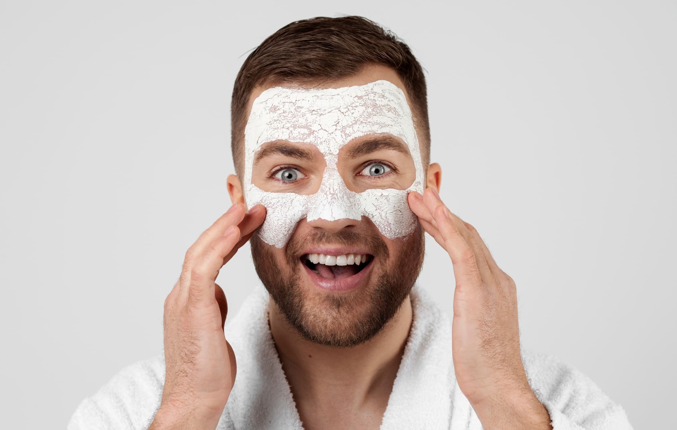 A smiling man wearing a bathrobe applying an exfoliating face mask, enjoying his skincare routine. His playful expression highlights the fun and relaxing side of self-care.