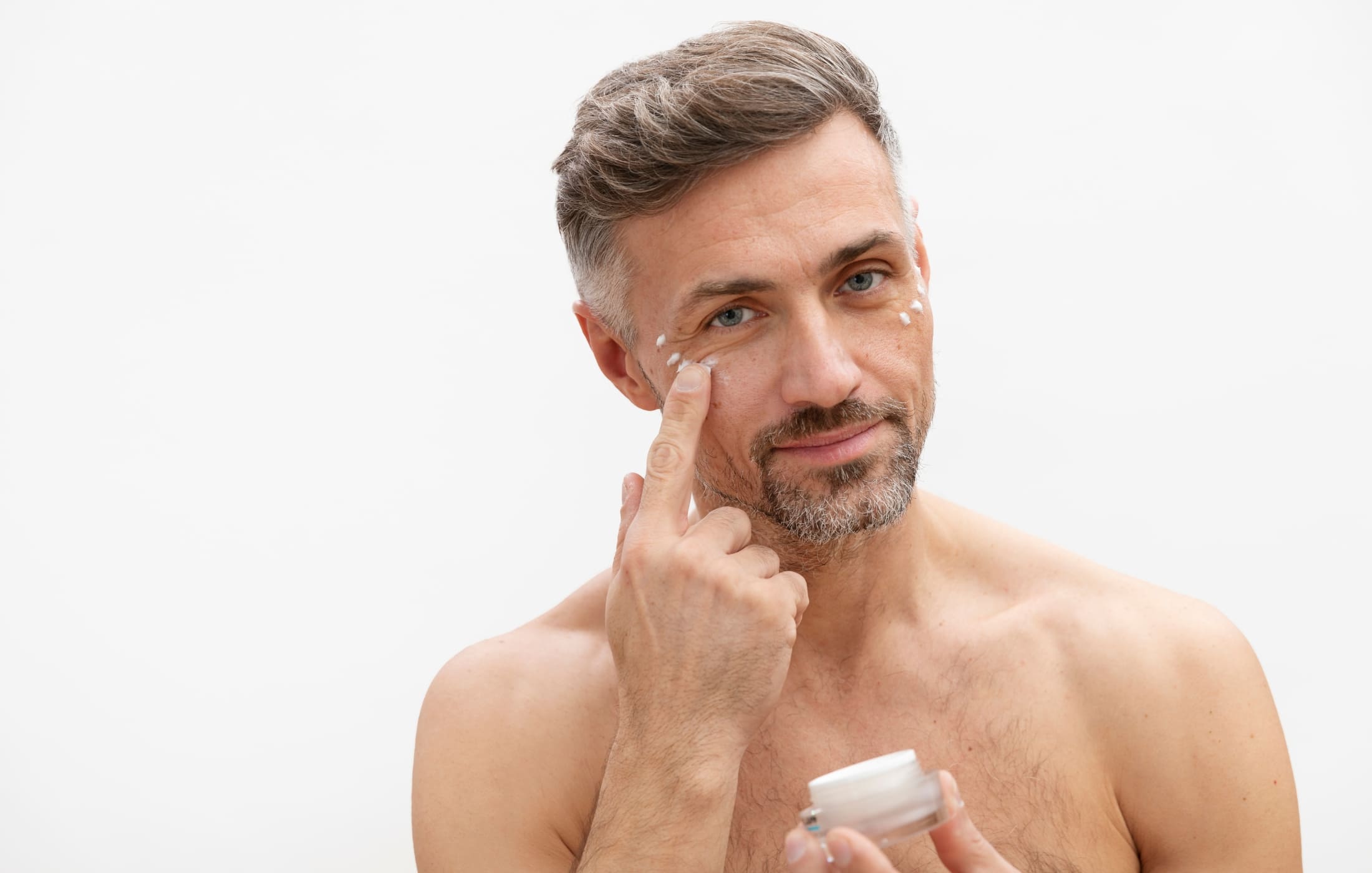 A close-up of a man with salt-and-pepper hair and a neatly groomed beard applying eye cream under his eyes, showcasing an important step in a men's skincare routine. He is gently dabbing small dots of cream around his eye area, smiling confidently as he cares for his skin. The minimal background keeps the focus on his face and the skincare process, emphasizing the importance of targeted treatments like eye cream in maintaining a youthful appearance.