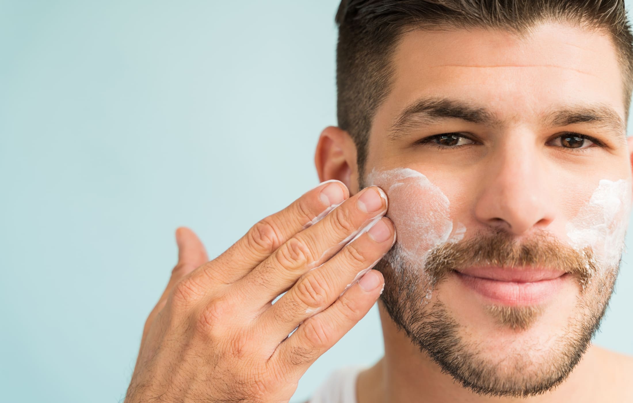 A close-up of a man with neatly groomed facial hair applying moisturizer to his face. He is gently massaging the cream into his skin, showcasing a key step in skincare tips for men. The light background highlights his clear, healthy complexion, emphasizing the importance of moisturizing in a daily skincare routine. His smile and relaxed expression suggest he’s enjoying the process, making this image a great example of how easy and beneficial men’s skincare can be.