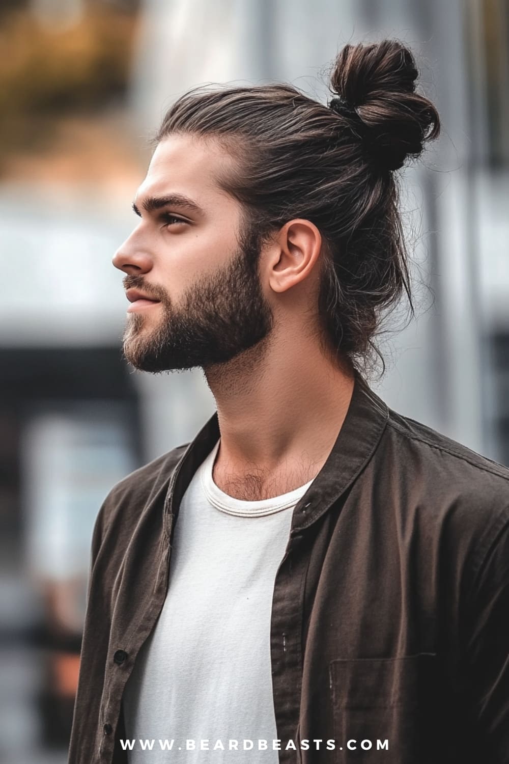 A man confidently sports a classic man bun, a popular choice among long hairstyles for men with thick hair. His thick hair is neatly pulled back into a high bun, showcasing the full volume and texture of his long locks. The look is complemented by a well-groomed beard, adding to his rugged yet polished appearance. He is dressed casually in a white t-shirt layered with an open dark jacket, exuding a relaxed and effortlessly cool vibe.