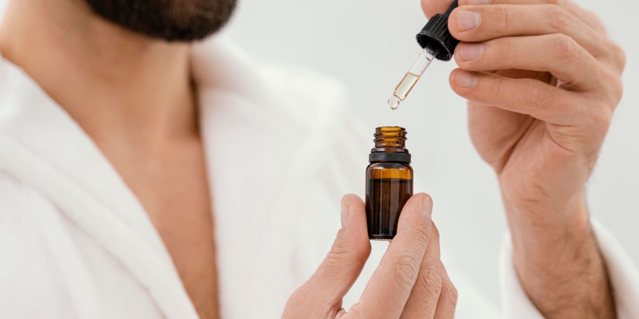 Man dispensing beard oil from a dropper bottle: Close-up of a bearded man in a white robe applying beard oil, highlighting the importance of beard grooming.