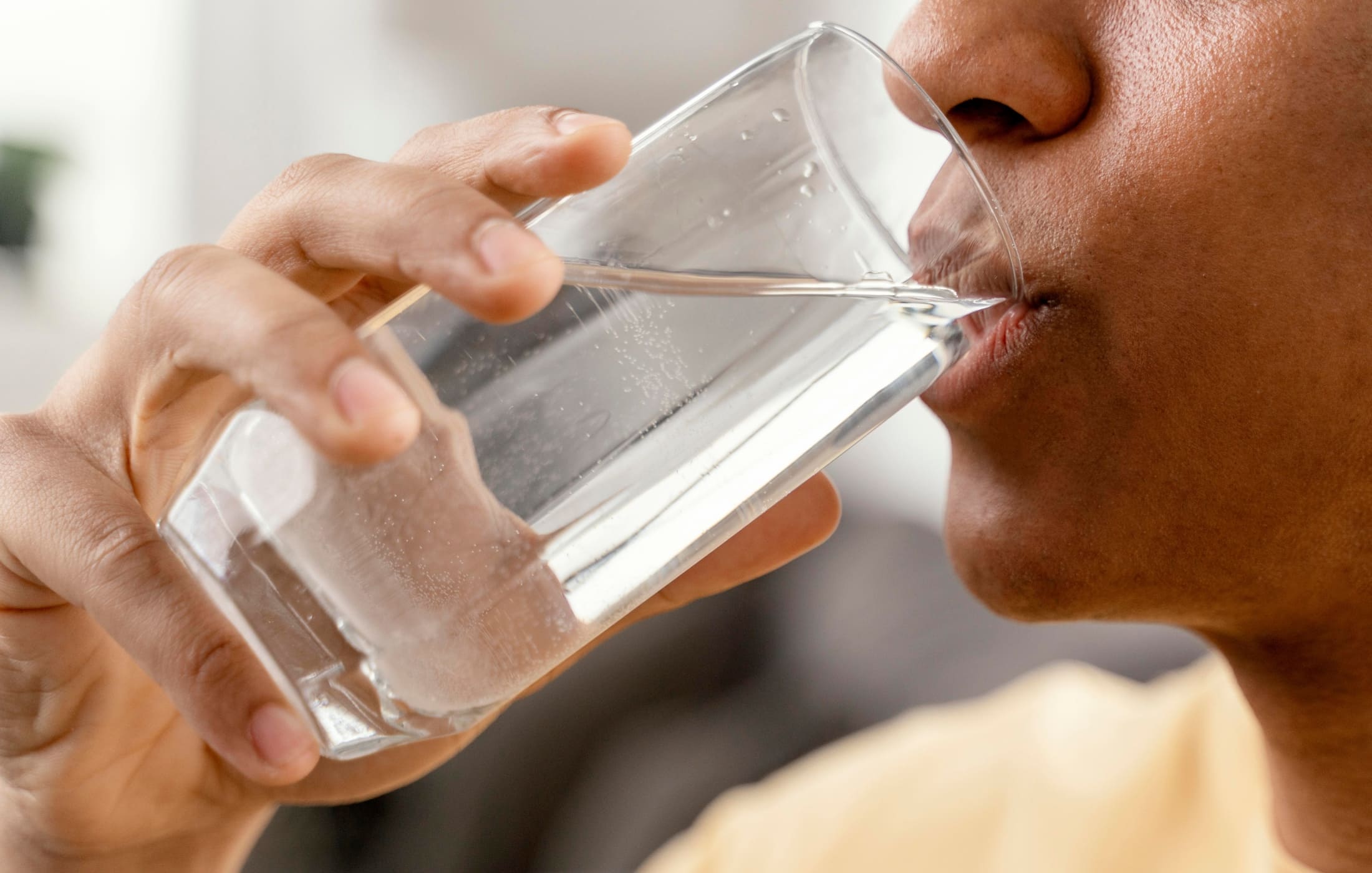 A close-up of a person drinking a glass of water, highlighting the importance of staying hydrated. The clear glass reflects the light, and you can see tiny bubbles clinging to the inside. The person’s hand is gently holding the glass, and their lips are just touching the rim as they take a sip.