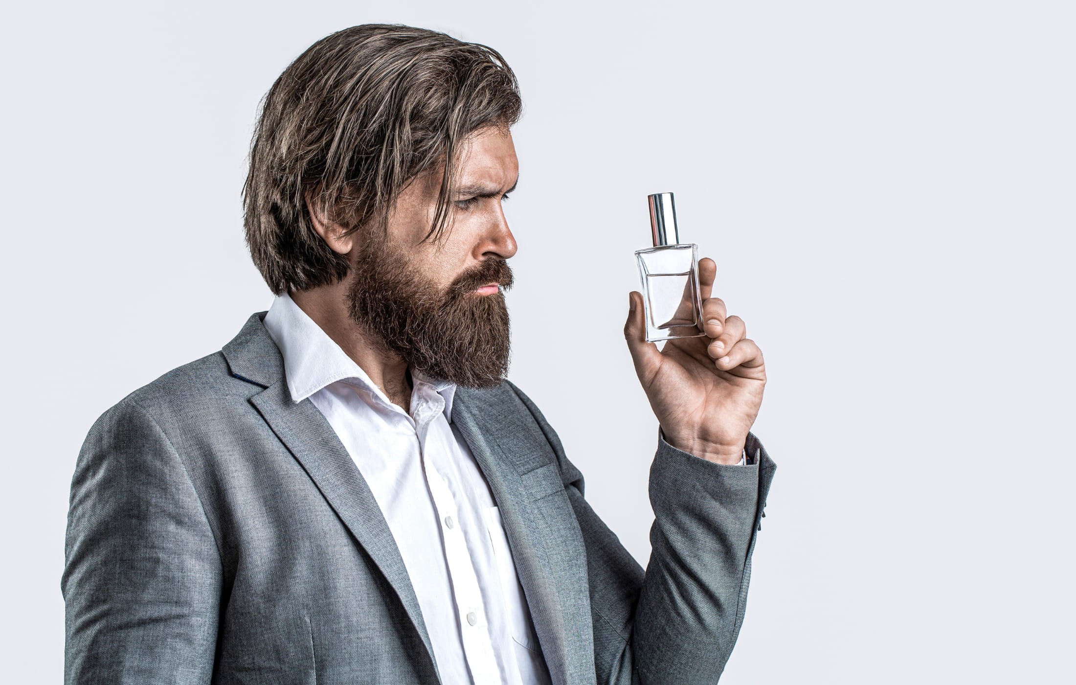 Bearded man in a gray suit thoughtfully examining a bottle of cologne, highlighting the importance of selecting the right fragrance for a polished and confident look.