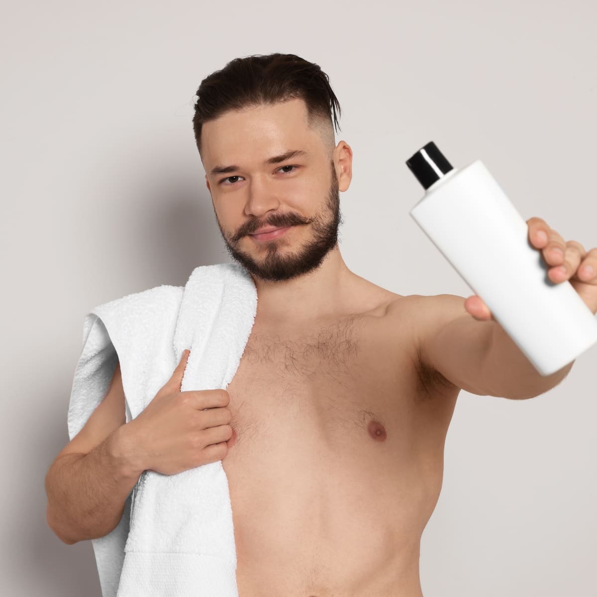 A young man with a neatly trimmed beard and mustache, and slicked-back hair, stands shirtless holding a white bottle of shower gel towards the camera. He has a white towel draped over one shoulder and a confident, friendly expression on his face. The background is a plain, light color, keeping the focus on the man and the grooming product he's showcasing.