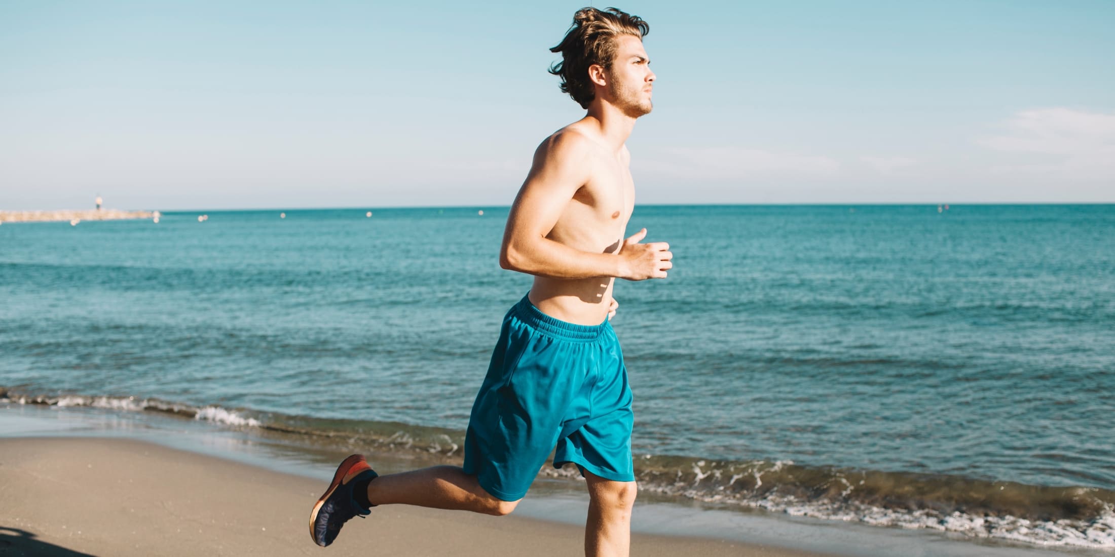 A shirtless man jogs along a sunny beach, practicing self-care for men by staying active and engaging in outdoor fitness. The calm ocean and clear sky in the background highlight the importance of exercise, relaxation, and mental well-being as part of a healthy lifestyle.