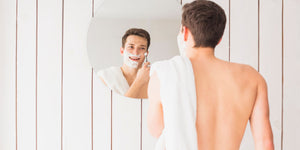  A young man smiling as he shaves in front of a mirror. He has shaving cream on his face and is wrapped in a white towel. His reflection shows his focus on learning how to get rid of 5 o'clock shadow