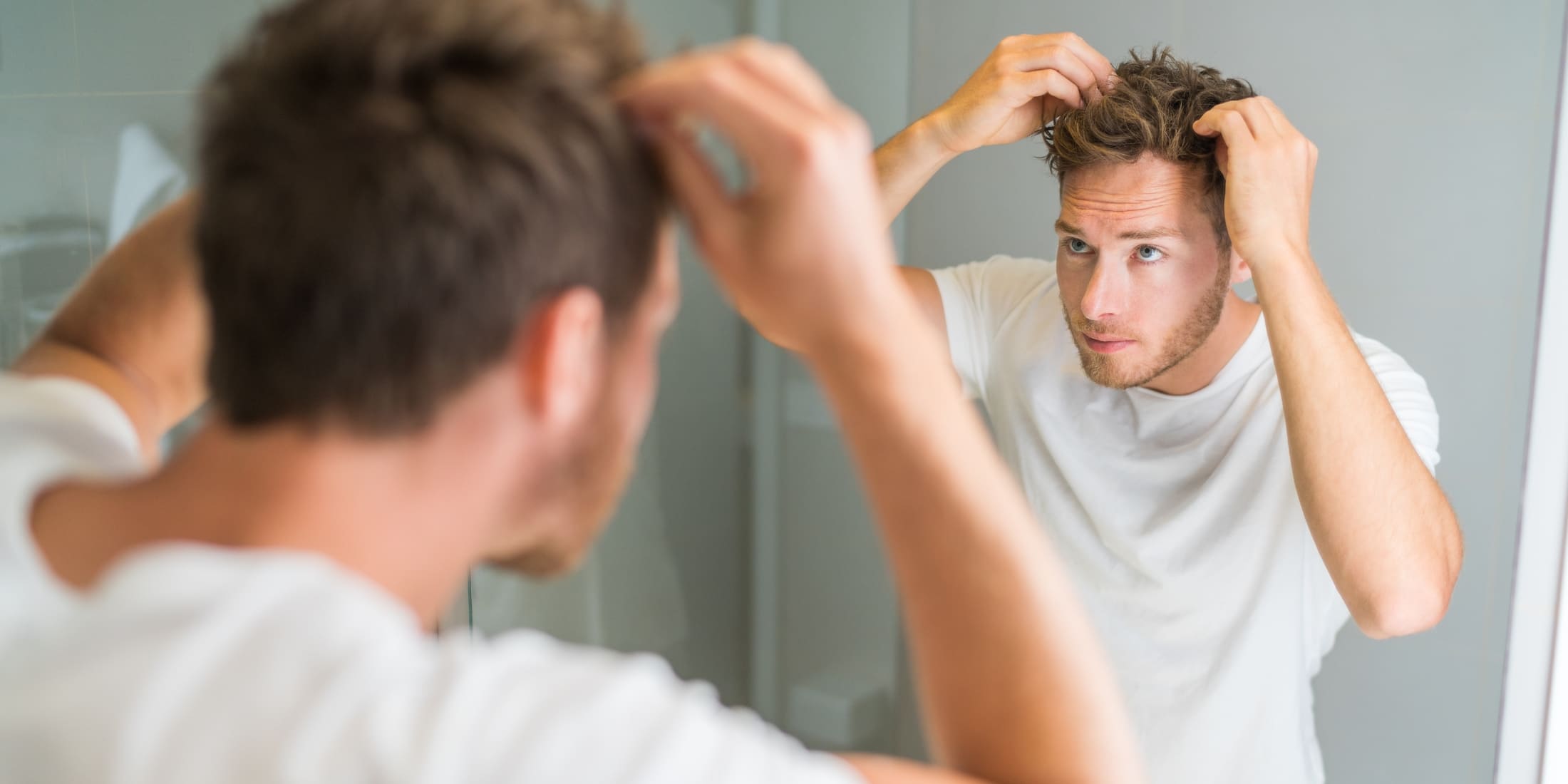 A man with a focused expression is standing in front of a bathroom mirror, learning how to use hair paste to style his short, textured hair. He is wearing a white T-shirt and using both hands to apply the paste evenly. The background is a clean, modern bathroom, reflecting his grooming routine.