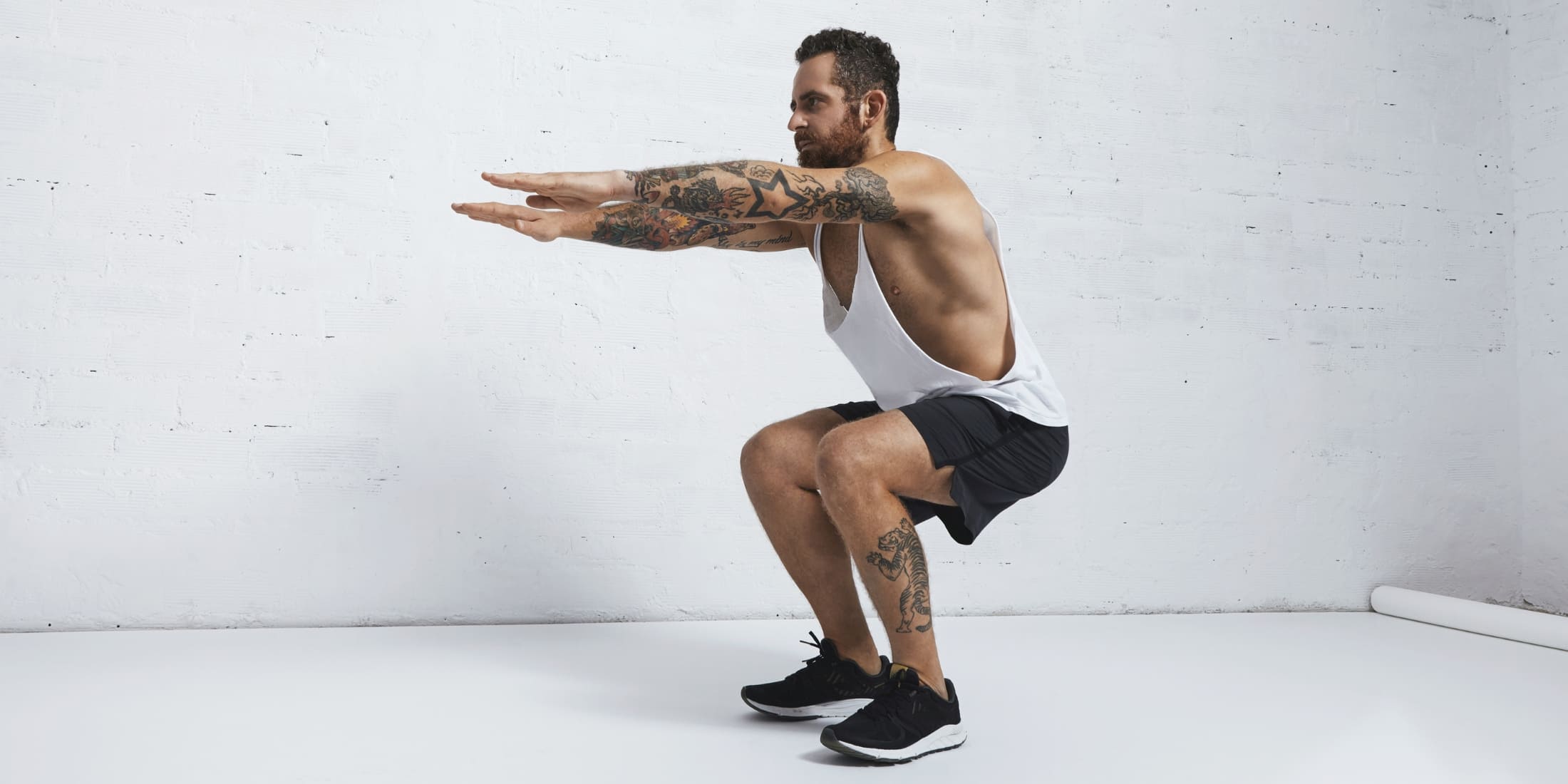 A tattooed man performing a bodyweight squat with arms extended in front. He is wearing a white tank top, black shorts, and black sneakers. This exercise targets the legs, glutes, and core, making it a fundamental part of a simple bodyweight workout or a beginner workout for men.