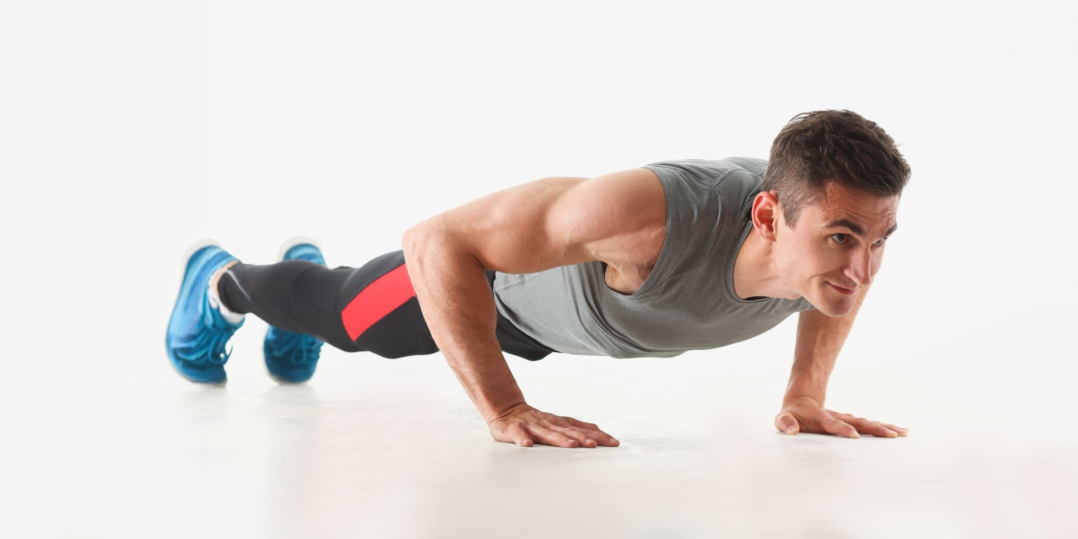 A fit man performing a push-up in a plank position. He is wearing a gray tank top, black leggings with red accents, and blue shoes. This bodyweight exercise targets the upper body and core, making it a great addition to a simple morning workout for men.