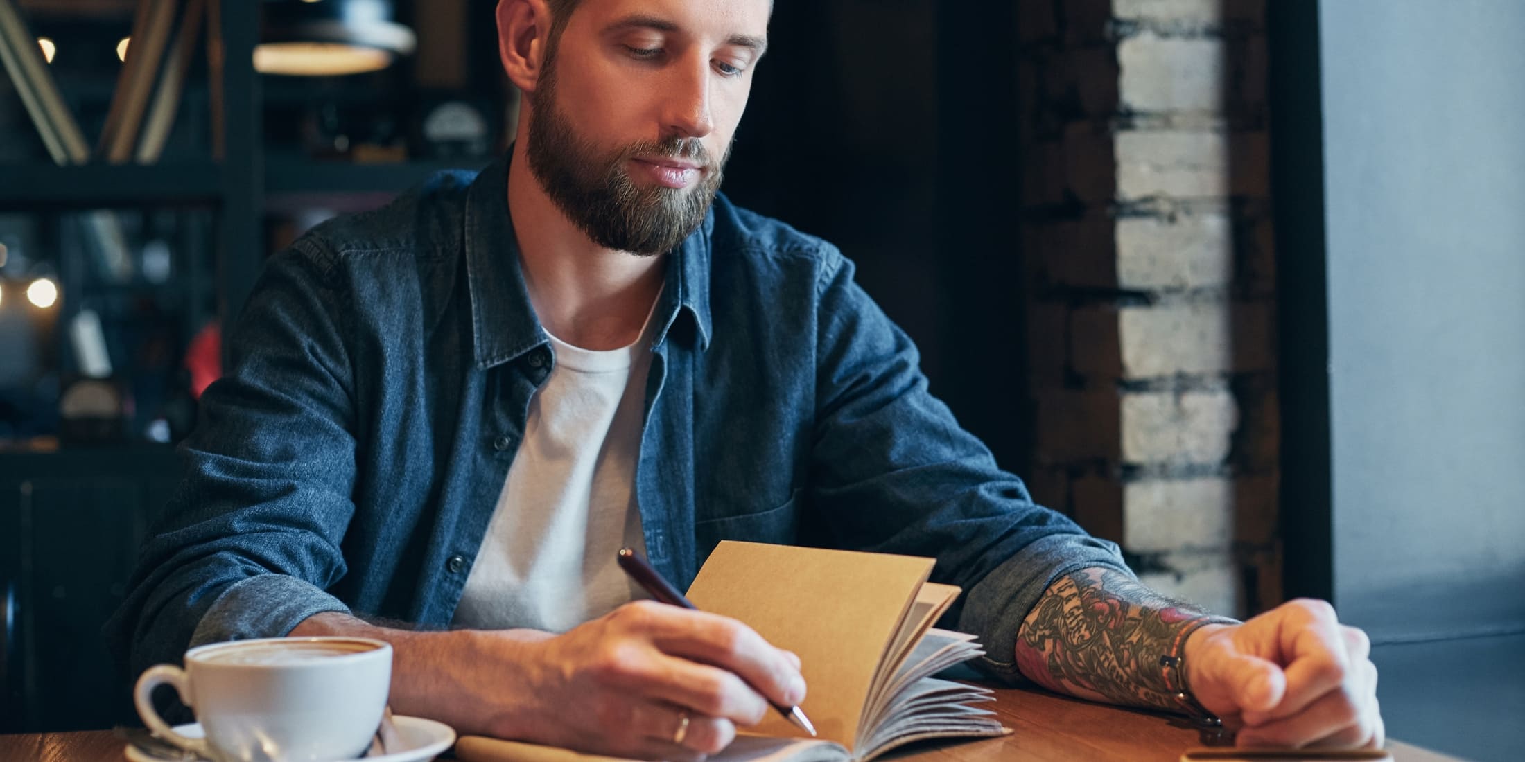 A man sitting at a table with a coffee, thoughtfully planning his day by writing in a journal.