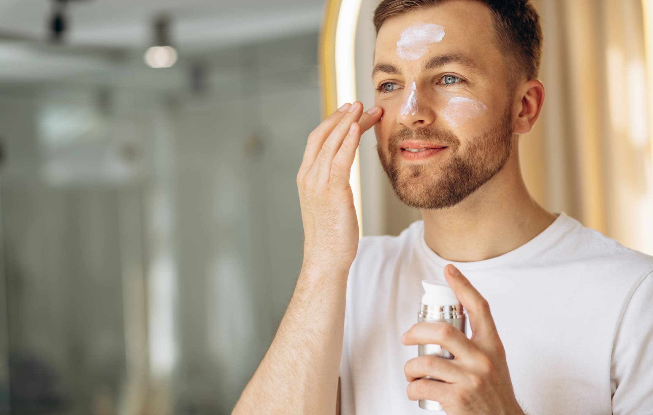 A man in a bright room applying skincare cream to his face, smiling as he practices his daily skincare routine. He holds a bottle of moisturizer while gently rubbing the cream into his skin, emphasizing the importance of self-care and healthy skin.