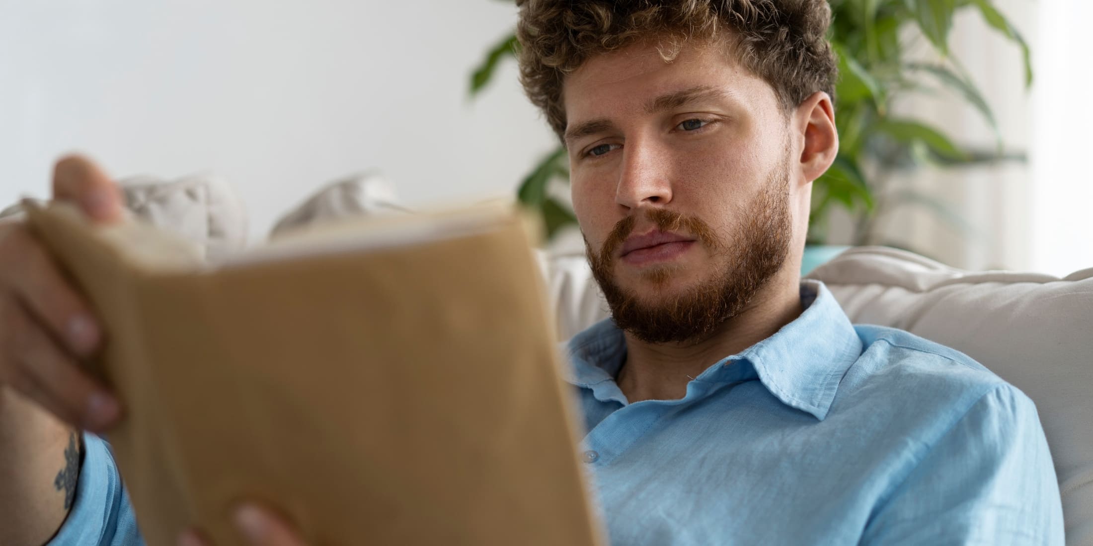 A man sitting comfortably on a couch, reading a book or mindfulness journal, focusing on self-reflection and relaxation, practicing mindfulness for men to improve mental clarity and emotional well-being.