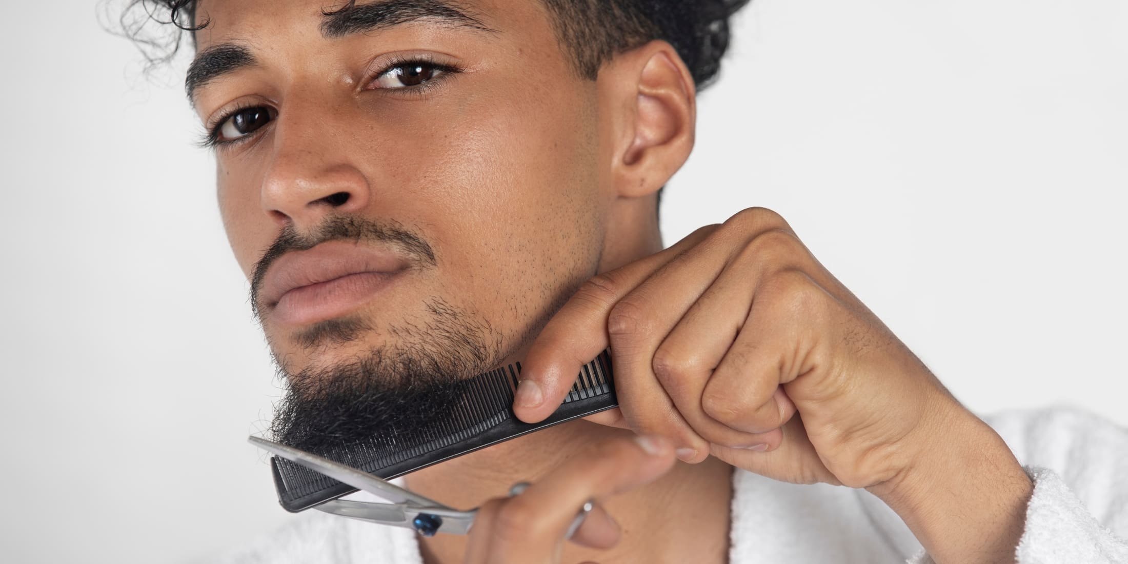 A man carefully trims his beard with scissors and a comb, demonstrating the importance of personal grooming as part of self-care for men. His focused expression highlights the significance of maintaining a well-groomed appearance for confidence and self-presentation.