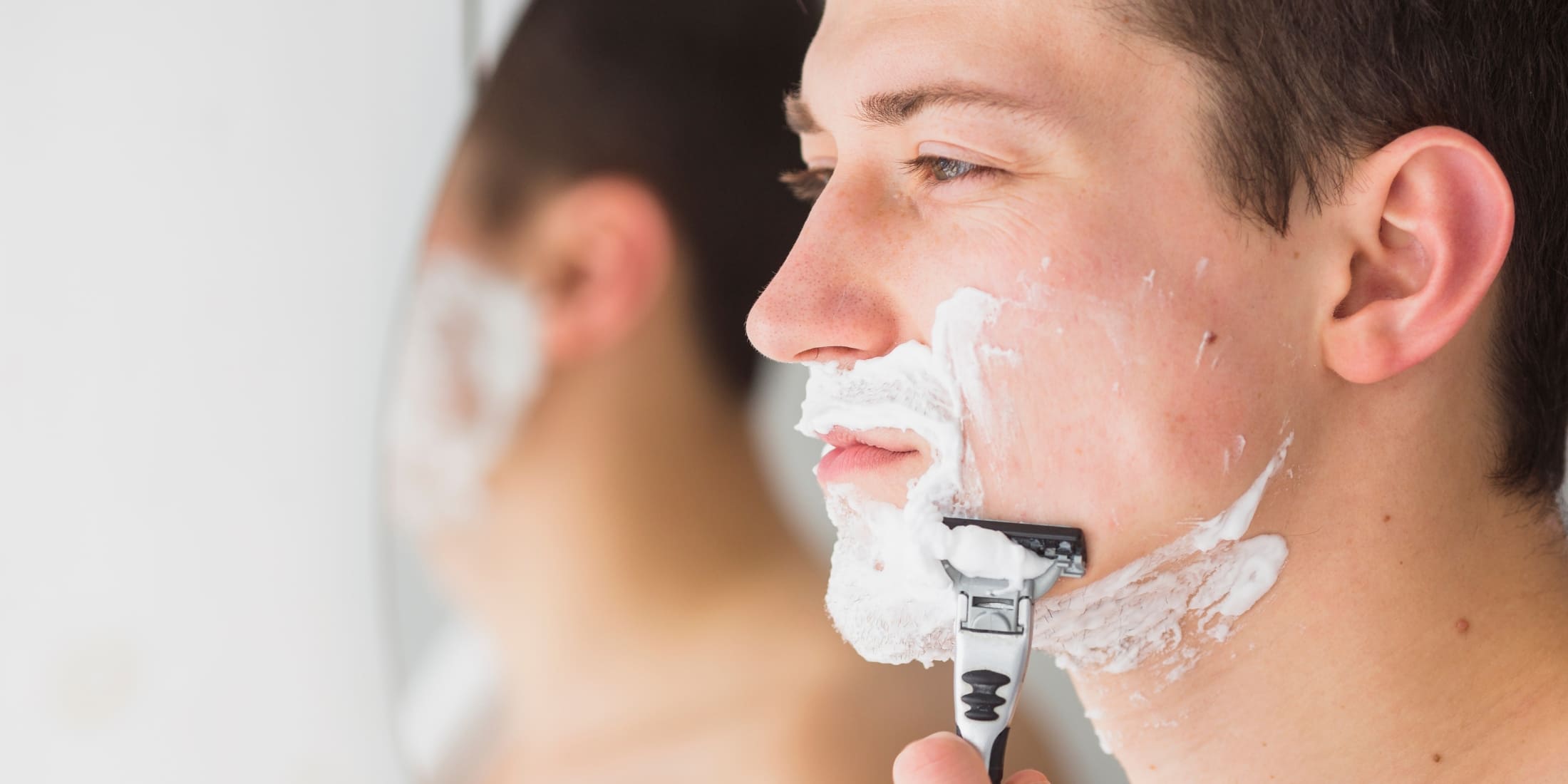 A close-up side view of a young man shaving his face. He has shaving cream applied generously on his cheeks, chin, and upper lip. He is using a razor to carefully shave his beard, aiming to get rid of his 5 o'clock shadow. The background shows his reflection in a mirror, emphasizing his focused expression and the precision of his shaving routine.