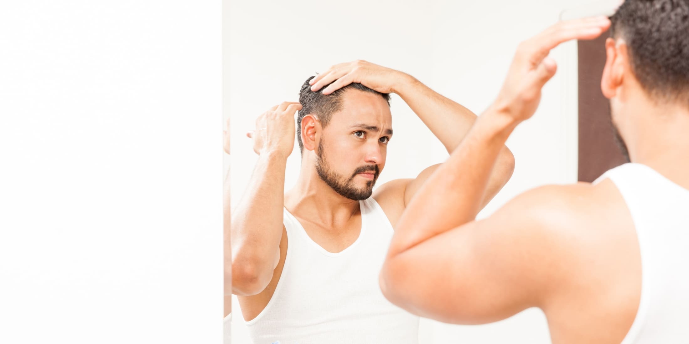 A man standing in front of a mirror, applying hair gel to style his hair. He is wearing a white tank top and appears to be focusing on achieving a polished look. This image illustrates what does hair gel do by showing its use in creating and maintaining a well-groomed hairstyle.