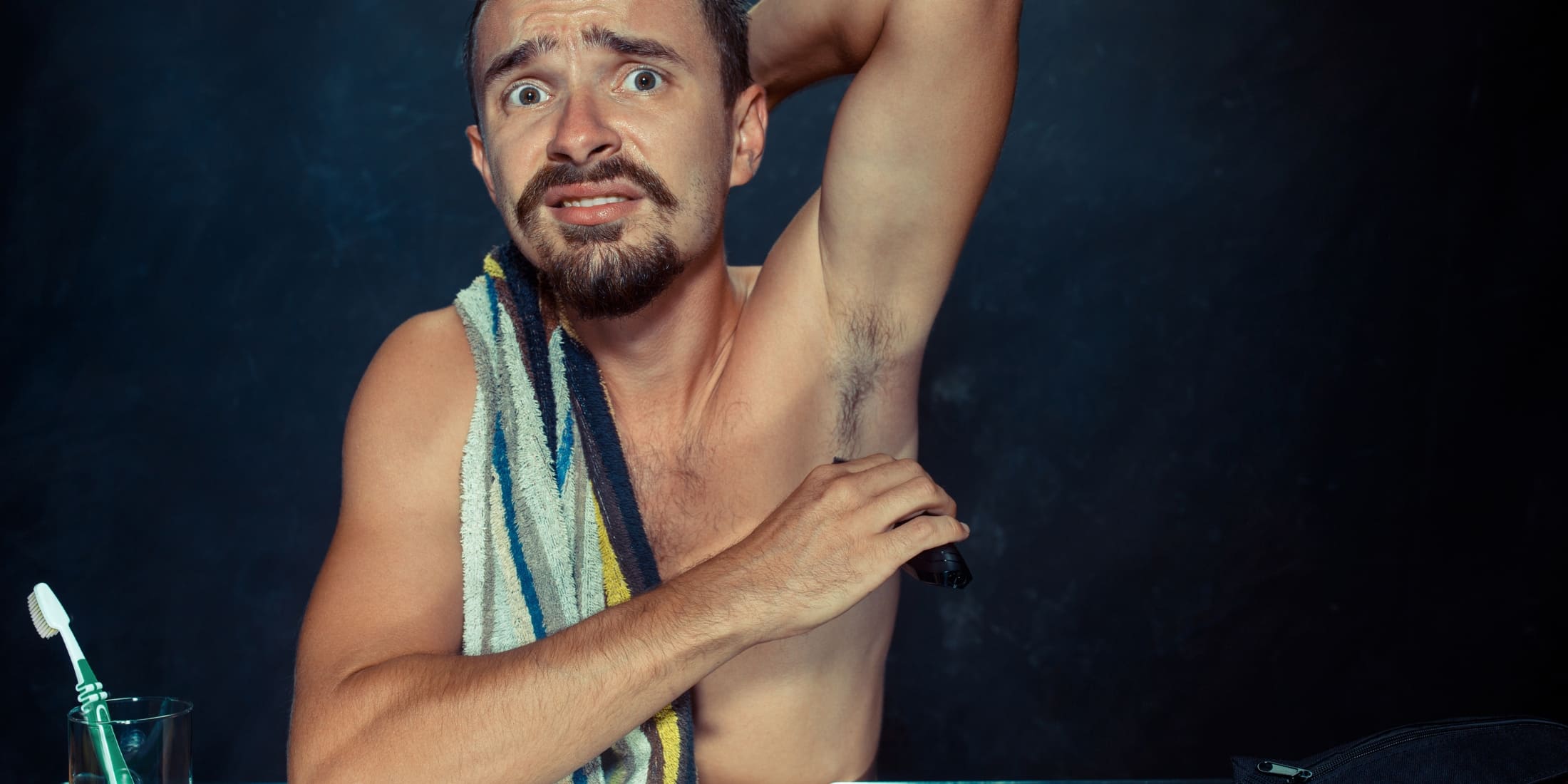 A man is seen in a bathroom setting, holding an electric trimmer to his armpit hair. He has a slightly anxious expression on his face, with wide eyes and raised eyebrows, suggesting uncertainty about the task. His left arm is raised to expose his armpit, while his right hand holds the trimmer. He has a towel draped over his right shoulder, and a toothbrush is visible in a glass on the counter in front of him. The background is dark, making the man and his actions the focal point of the image.