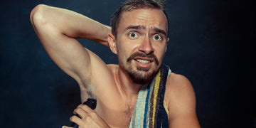 A man with a concerned expression is using an electric trimmer to trim his armpit hair. He has a towel over his shoulder and his arm raised to expose his armpit, demonstrating how to trim armpit hair.