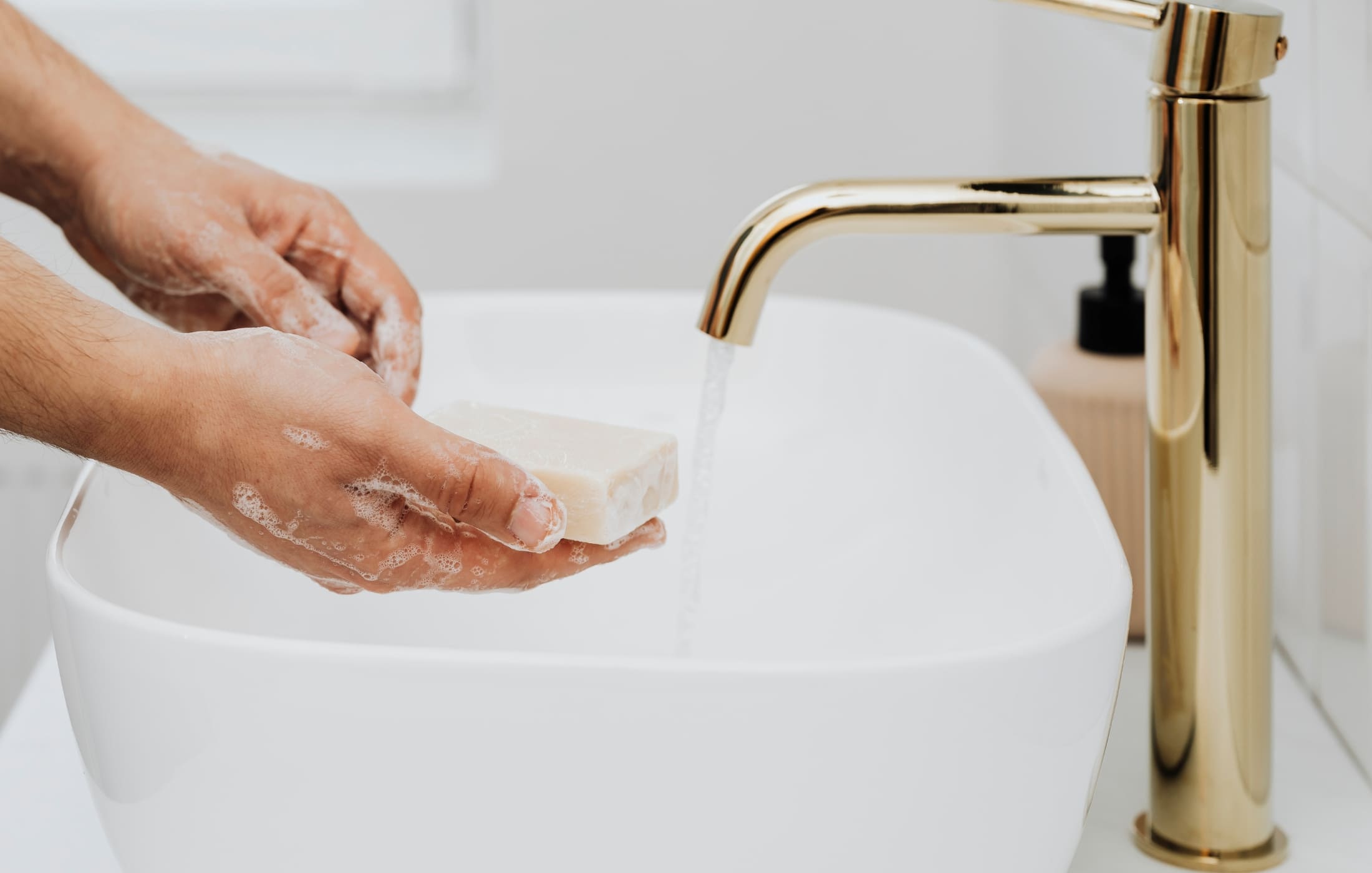 A close-up of a person washing their hands with a bar of soap under running water from a modern, gold-colored faucet. The person's hands are lathered with soap, creating a rich foam as they clean their hands. The bright white sink and the sleek faucet add a touch of elegance to the scene, emphasizing cleanliness and the importance of hygiene in daily routines. The focus on the hands and the soap creates a simple yet impactful image of a common, everyday practice.