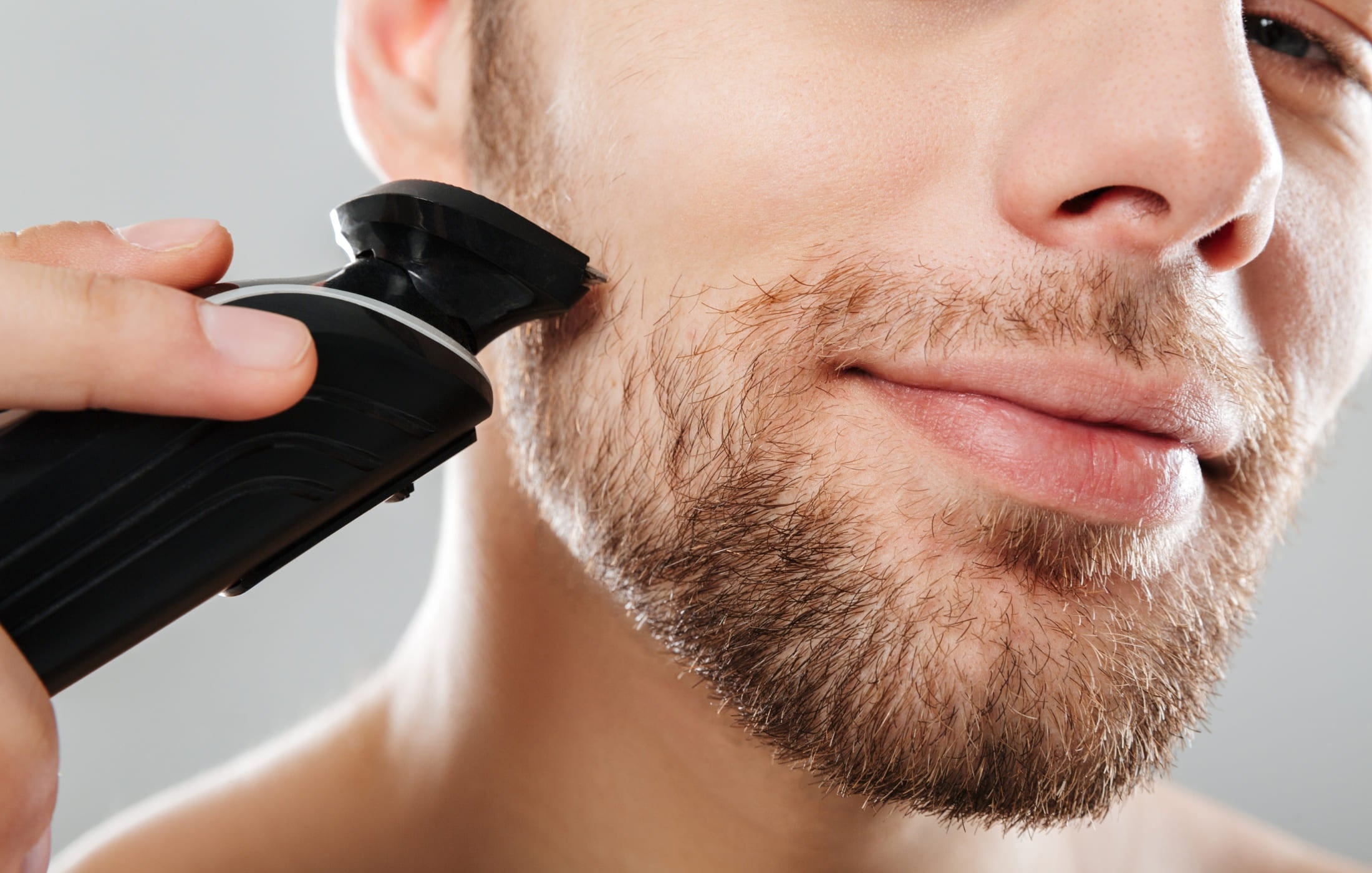 Close-up of a man using an electric trimmer to shape his beard, showing the precision and ease of grooming. The man has a light smile as he carefully trims his facial hair.