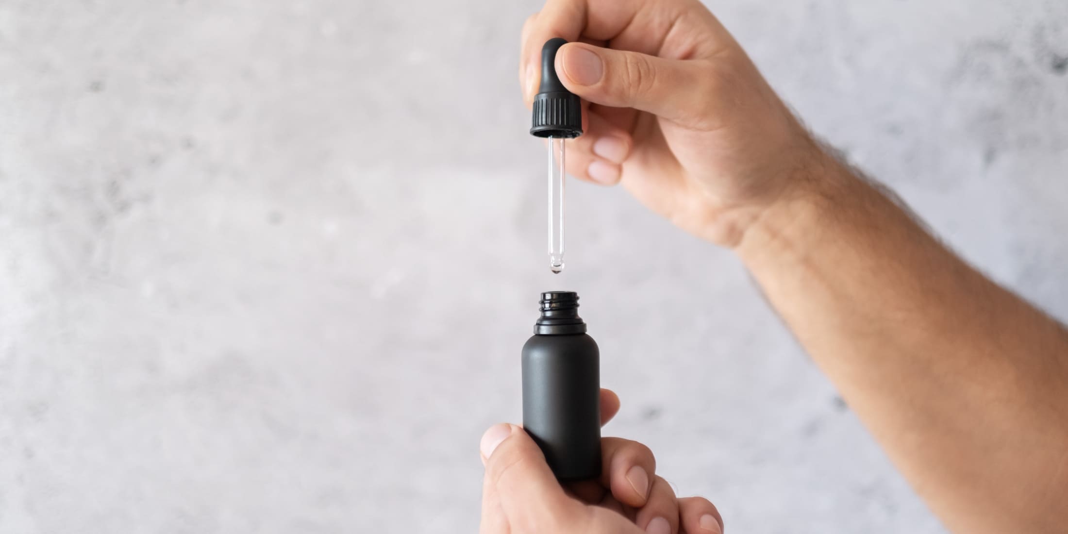 A person's hand holding a black dropper bottle, with a dropper dispensing a single drop of beard oil, against a blurred light grey background.