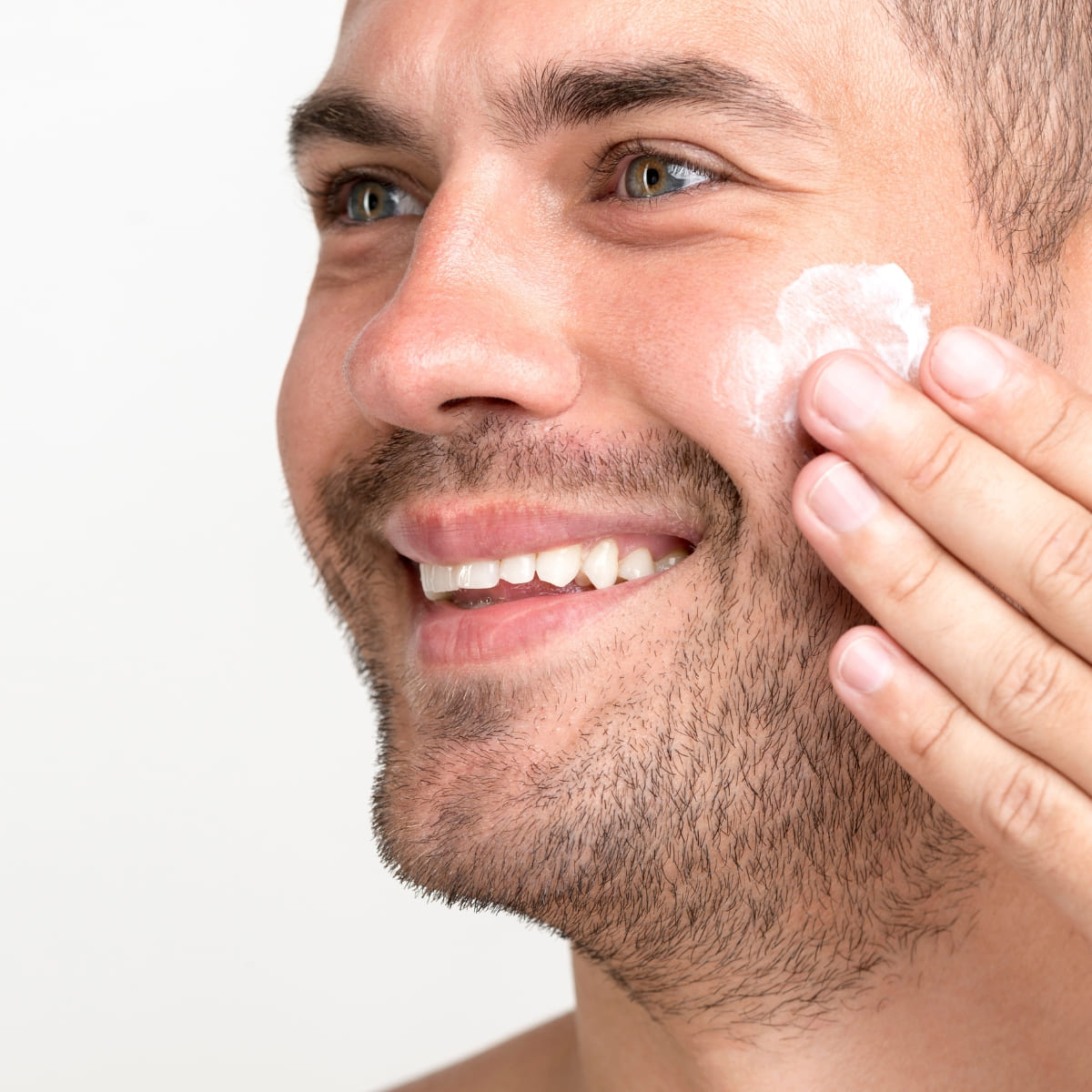 A close-up of a smiling man with a light beard and bright eyes applying moisturizer to his cheek. His hand is gently rubbing in the cream, showcasing a healthy skincare routine. The background is plain and light, keeping the focus on the man’s glowing skin and his grooming practice.