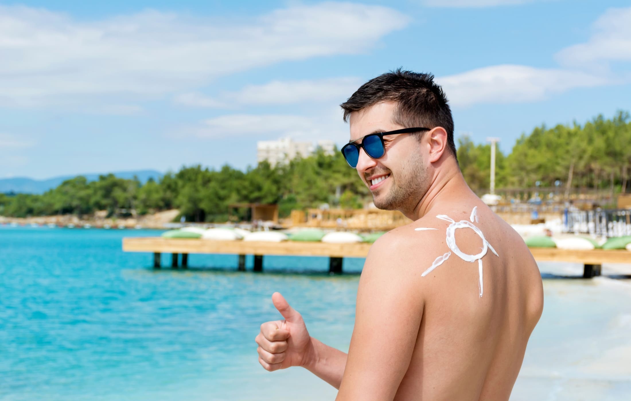 A smiling man at the beach gives a thumbs-up while showing off a sun drawn on his back with sunscreen. He wears sunglasses and enjoys the sunny weather, demonstrating the importance of sun protection in his men’s skincare routine.